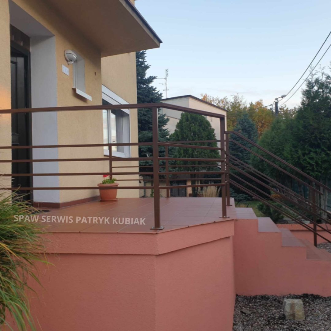 Zewnętrzna, brązowa balustrada metalowa z poziomych prętów, zabezpieczająca taras i schody wejściowe do domu, z doniczką z czerwoną pelargonią na tarasie.