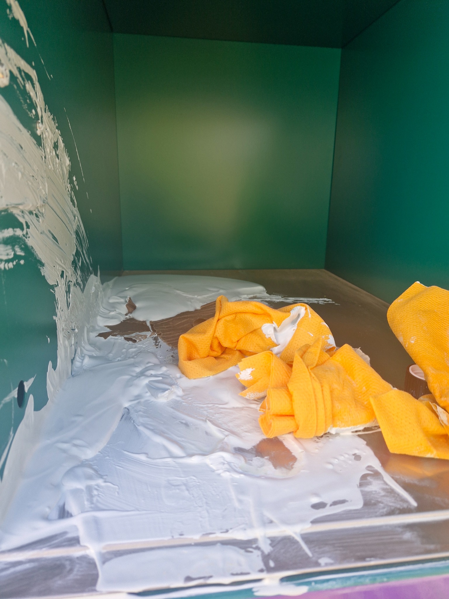 Świeżo pomalowane wnętrze szafki z zielonymi ściankami, z resztkami białej farby i żółtą ścierką leżącą na mokrej powierzchni.