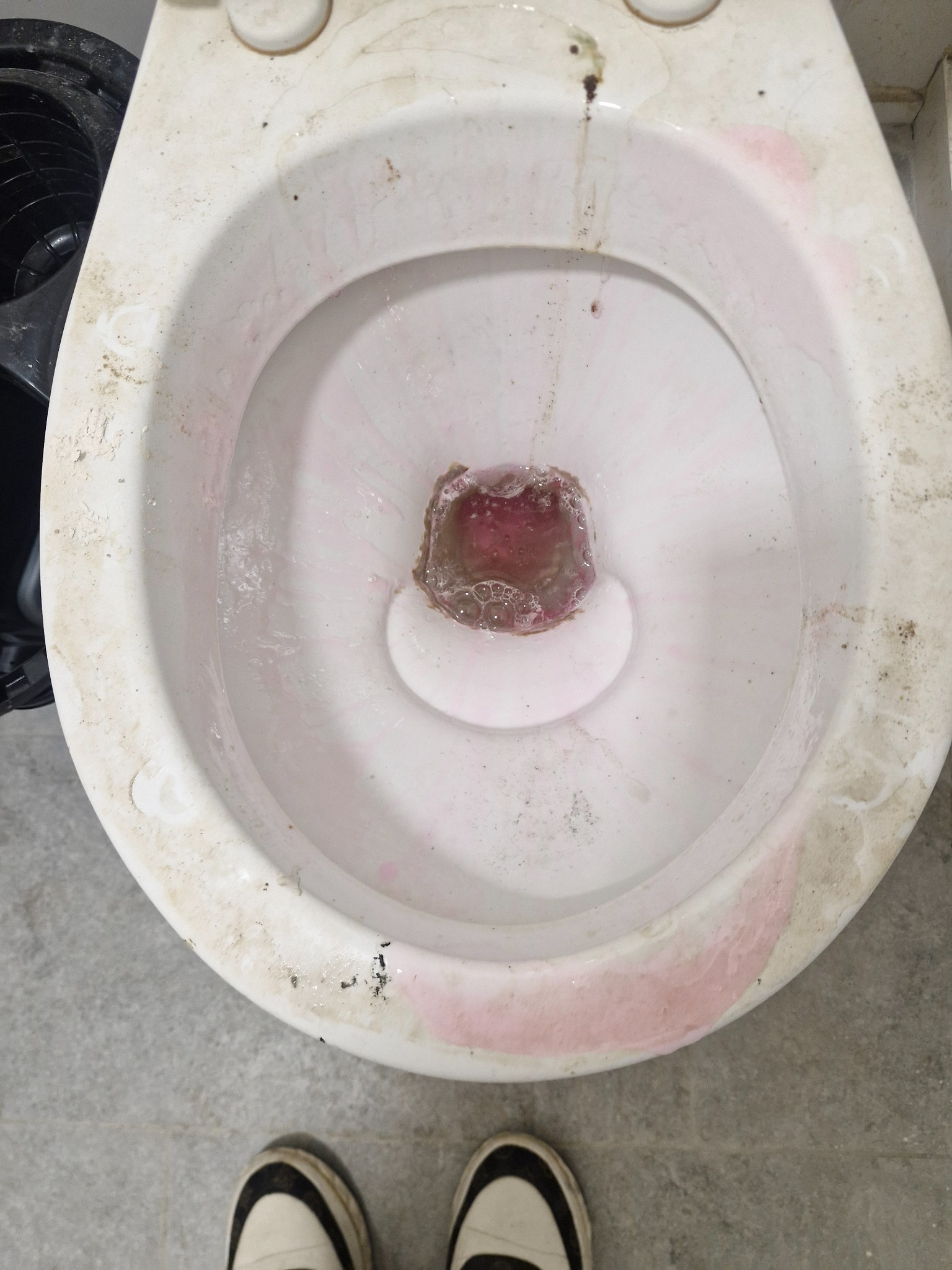 Zaniedbana toaleta z widocznymi zabrudzeniami i osadami, wypełniona wodą z różowym barwnikiem. U dołu kadru fragment butów osoby fotografującej.
