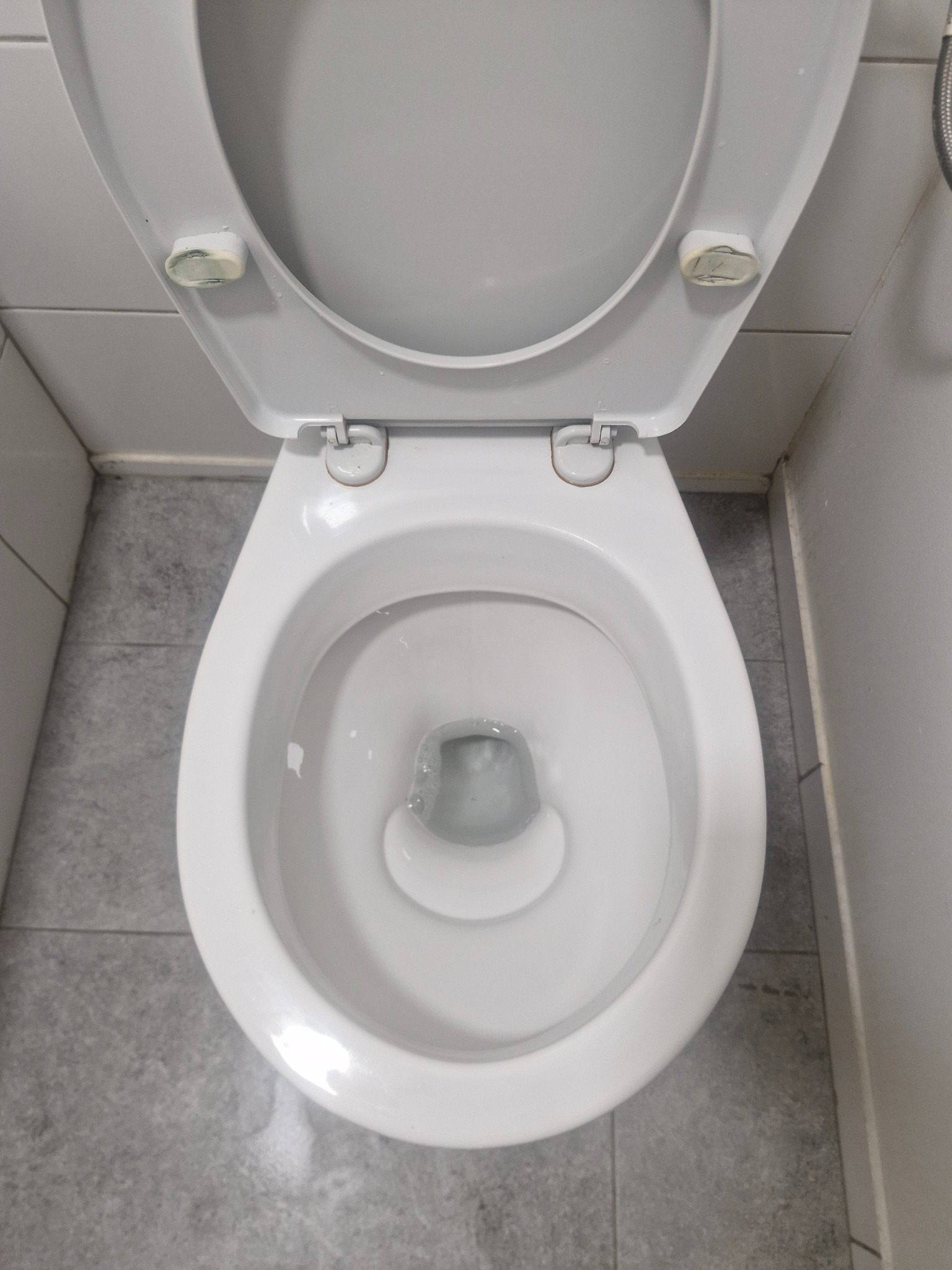 Biała, otwarta muszla klozetowa z wodą, widziana z góry na tle szarych płytek. Widoczne elementy mocujące klapę. Minimalistyczne ujęcie.