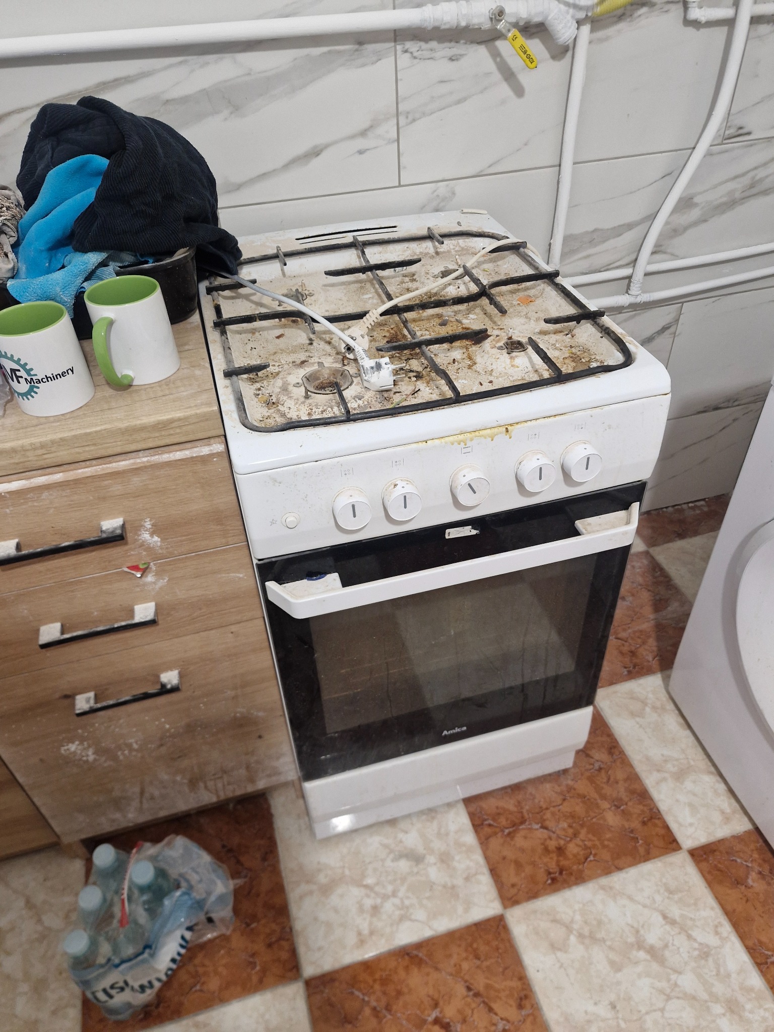 Bardzo zabrudzona, biała kuchenka gazowa Amica w kuchni z marmurowymi płytkami. Obok szafka z plamami i butelki wody. Widoczne rury gazowe.