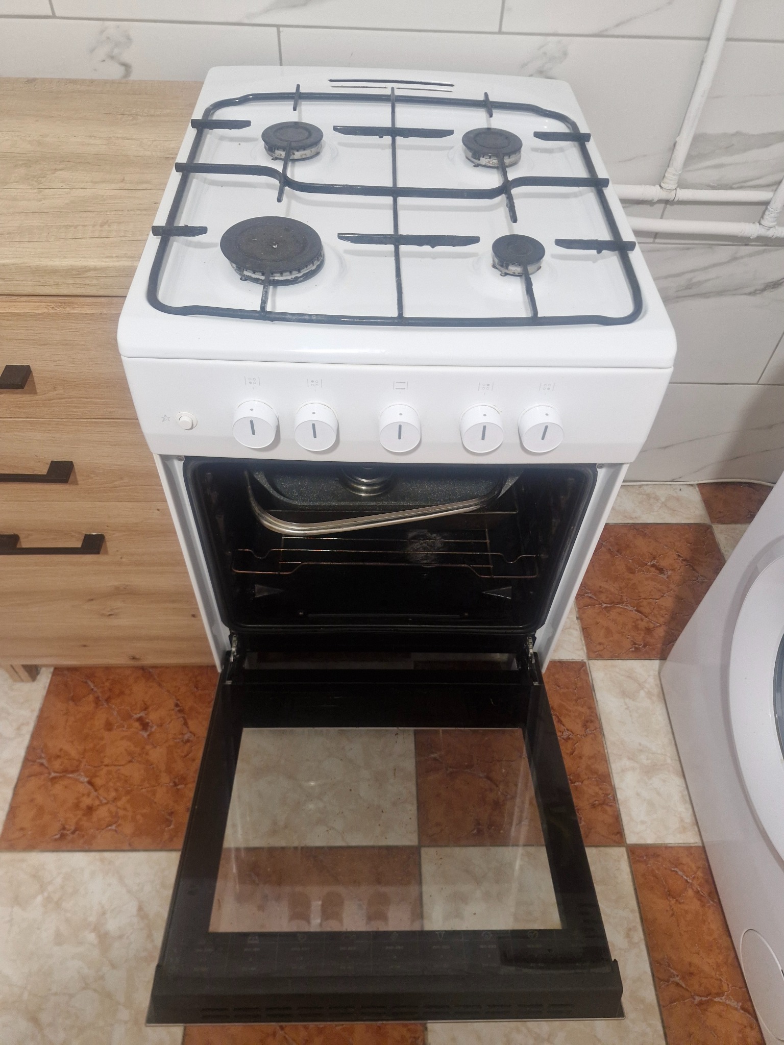 Biała kuchenka gazowa z otwartym piekarnikiem, widok z góry. Płyta z czterema palnikami, wnętrze piekarnika z blachą. Obok szafka i pralka. Płytki podłogowe w brązowo-beżową kratę.