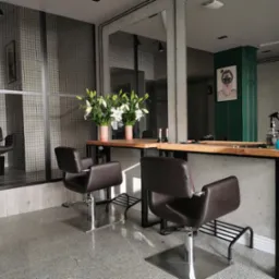 Nowoczesne stanowisko fryzjerskie z dwoma skórzanymi fotelami, drewnianym blatem i kwiatami w minimalistycznych doniczkach na tle ściany z metalowej siatki i lustra. Na blacie widoczne akcesoria...