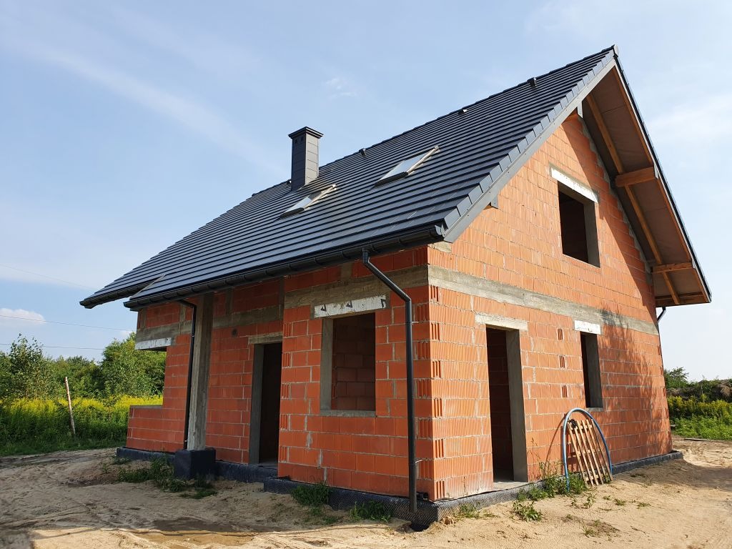 Surowy, dwukondygnacyjny budynek z czerwonej cegły z ciemnoszarą dachówką i kominem, widoczne otwory okienne i drzwiowe, w tle zieleń.