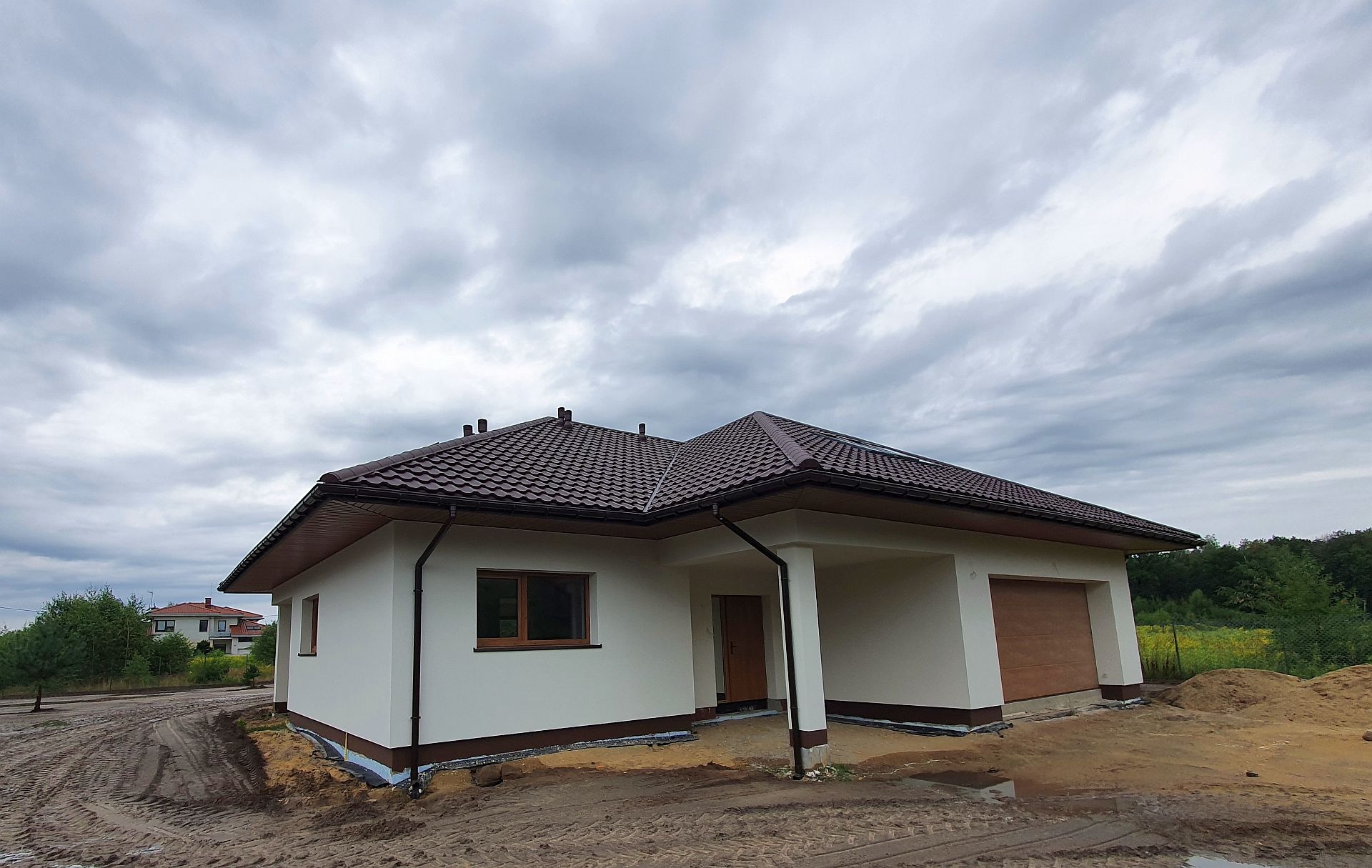 Niedawno ukończony parterowy dom z brązową dachówką, beżową elewacją i garażem w bryle, otoczony piaskiem i śladami opon, z widoczną izolacją przeciwwilgociową fundamentów.