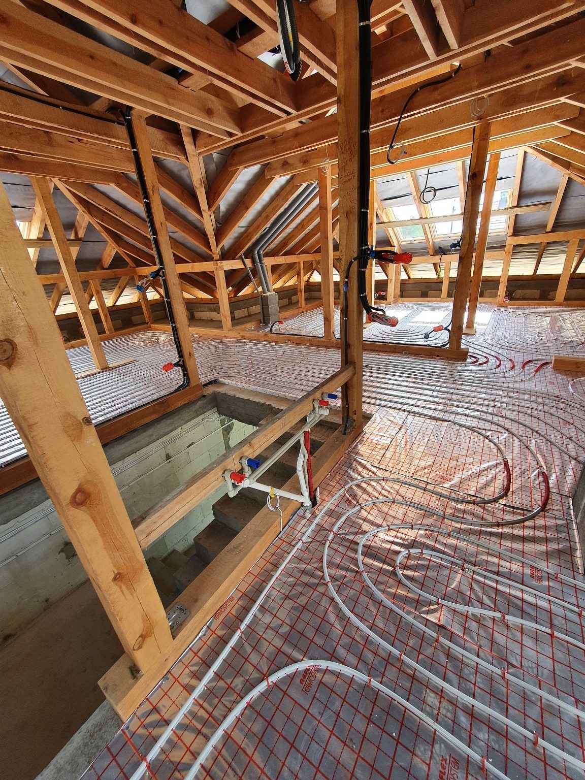 Poddasze w trakcie montażu ogrzewania podłogowego: białe rury ułożone na folii z czerwoną siatką, widoczna drewniana konstrukcja dachu i instalacje.