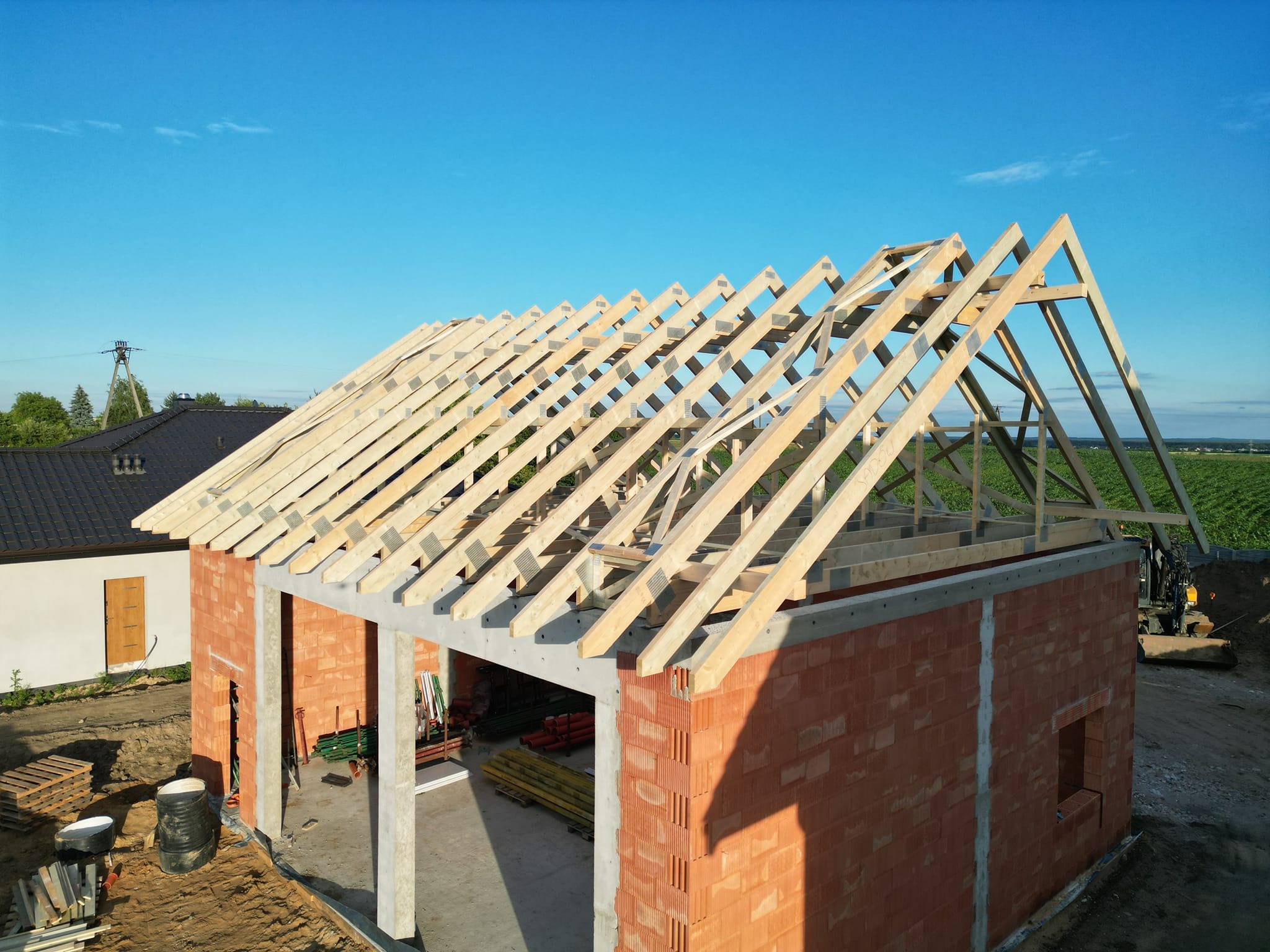 Wznoszenie ścian z czerwonej cegły ceramicznej i montaż drewnianej konstrukcji dachu na betonowym wieńcu, w tle zielone pole uprawne.