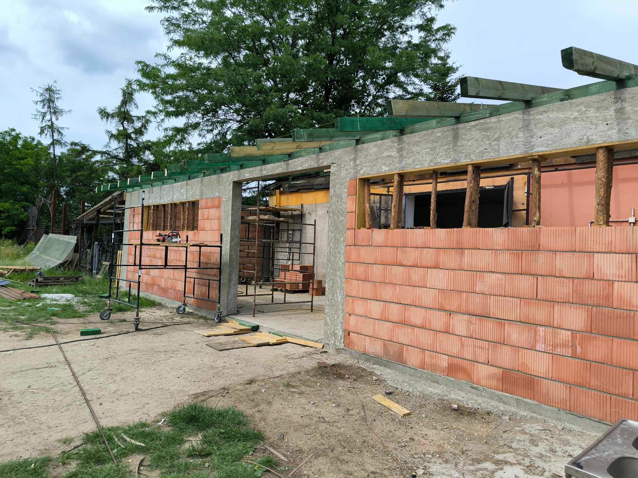 Wznoszenie ścian domu murowanego z czerwonej cegły, widoczne rusztowania, elementy drewniane konstrukcji dachu i teren budowy w tle.