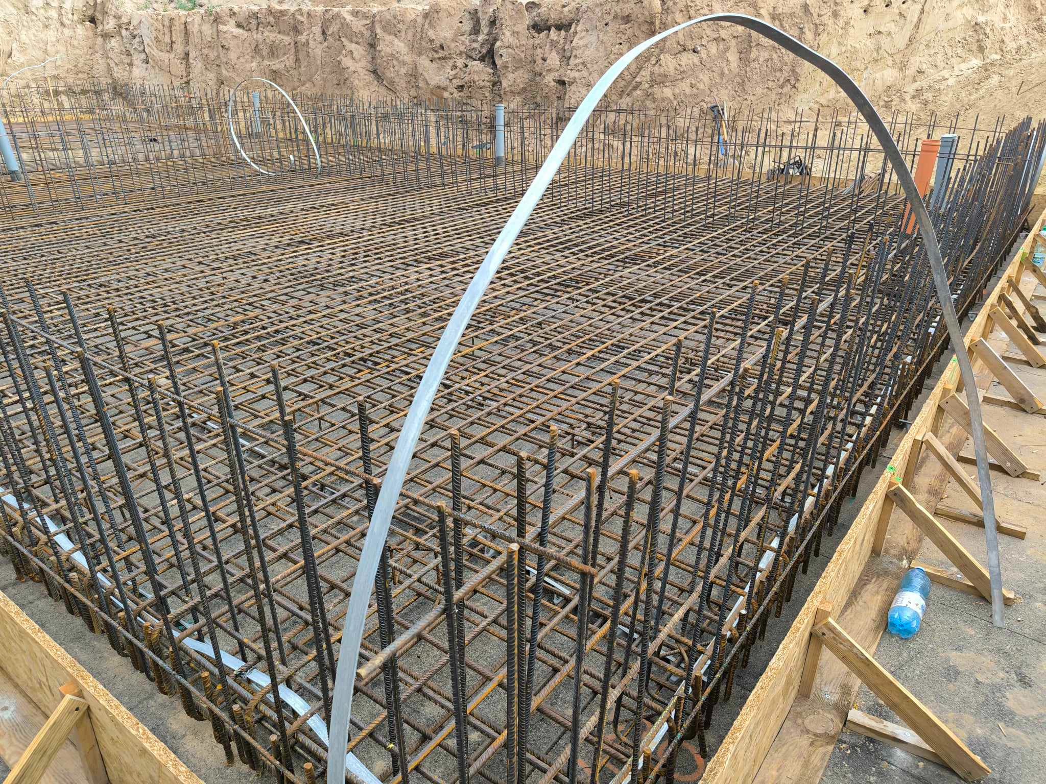 Zbrojenie płyty fundamentowej w wykopie, widoczne drewniane szalunki i stalowe pręty tworzące siatkę, u góry widoczny grunt.