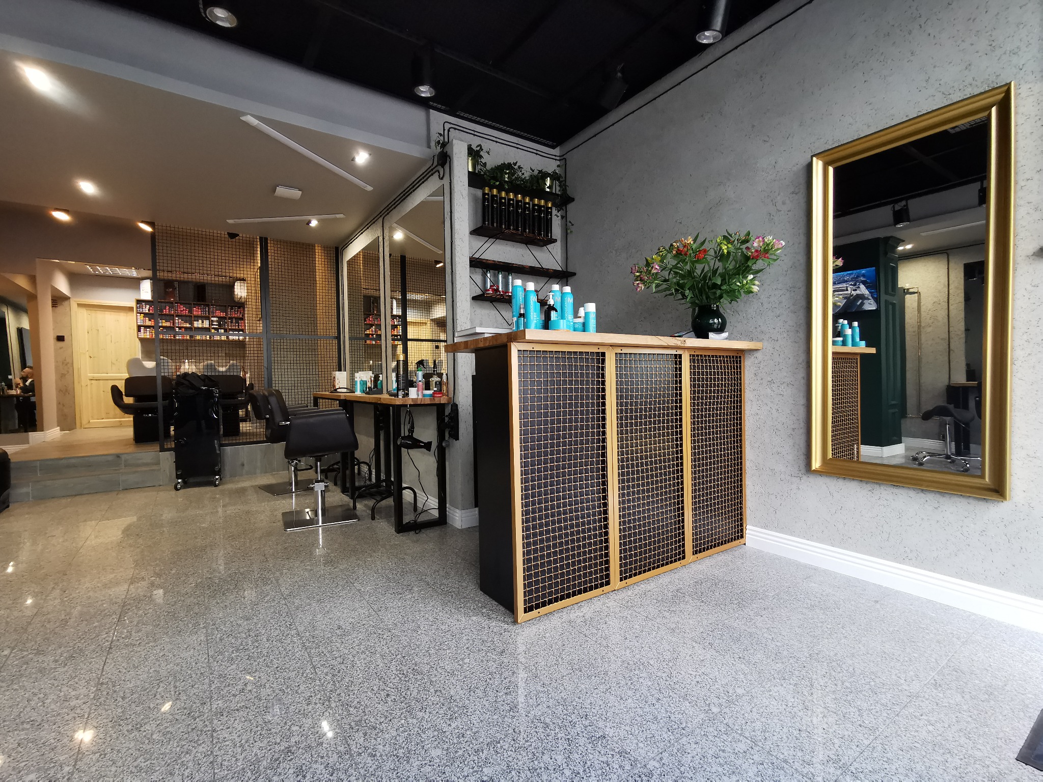 Nowoczesne wnętrze salonu fryzjerskiego z szarymi ścianami, marmurową podłogą, złotym lustrem i recepcją z kratką oraz stanowiskami fryzjerskimi w tle.