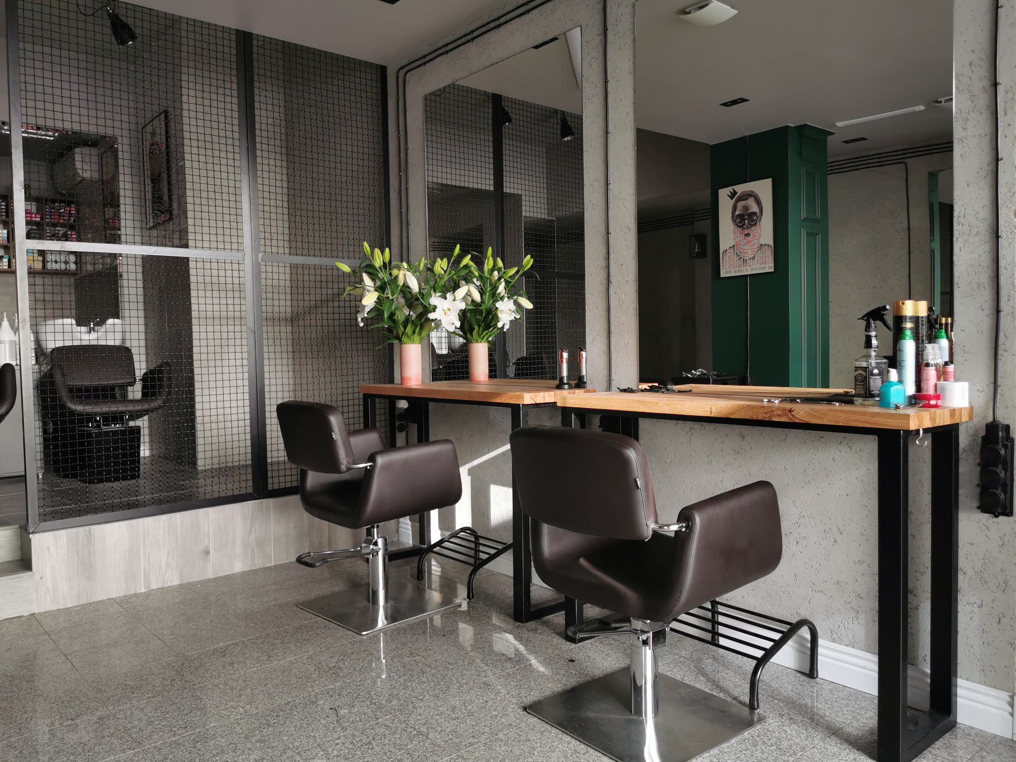 Nowoczesne stanowisko fryzjerskie z dwoma skórzanymi fotelami, drewnianym blatem i kwiatami w minimalistycznych doniczkach na tle ściany z metalowej siatki i lustra. Na blacie widoczne akcesoria...