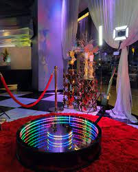 Okrągła platforma z lustrzanym blatem i wielokolorowym oświetleniem LED na czerwonym dywanie, część fotobudki na imprezie, w tle dekoracje i białe zasłony.