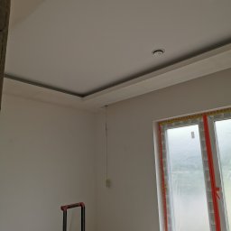 Piotr Modzelewski - Wnętrze pokoju w trakcie wykańczania: pomalowane ściany na biało, zamontowane oświetlenie sufitowe, okno zabezpieczone folią ochronną, widoczny fragment drabiny malarskiej.