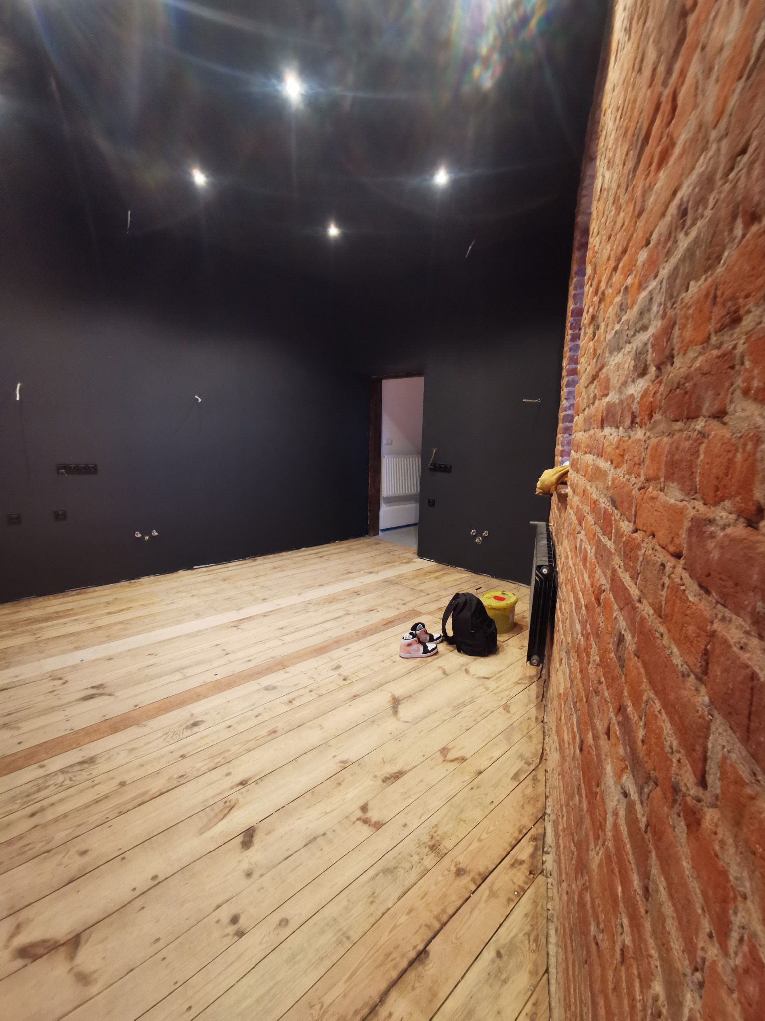 Pokój w trakcie remontu: czarne ściany, surowa drewniana podłoga, odsłonięta ceglana ściana, widoczne puszki elektryczne, torba i buty sportowe przy grzejniku.