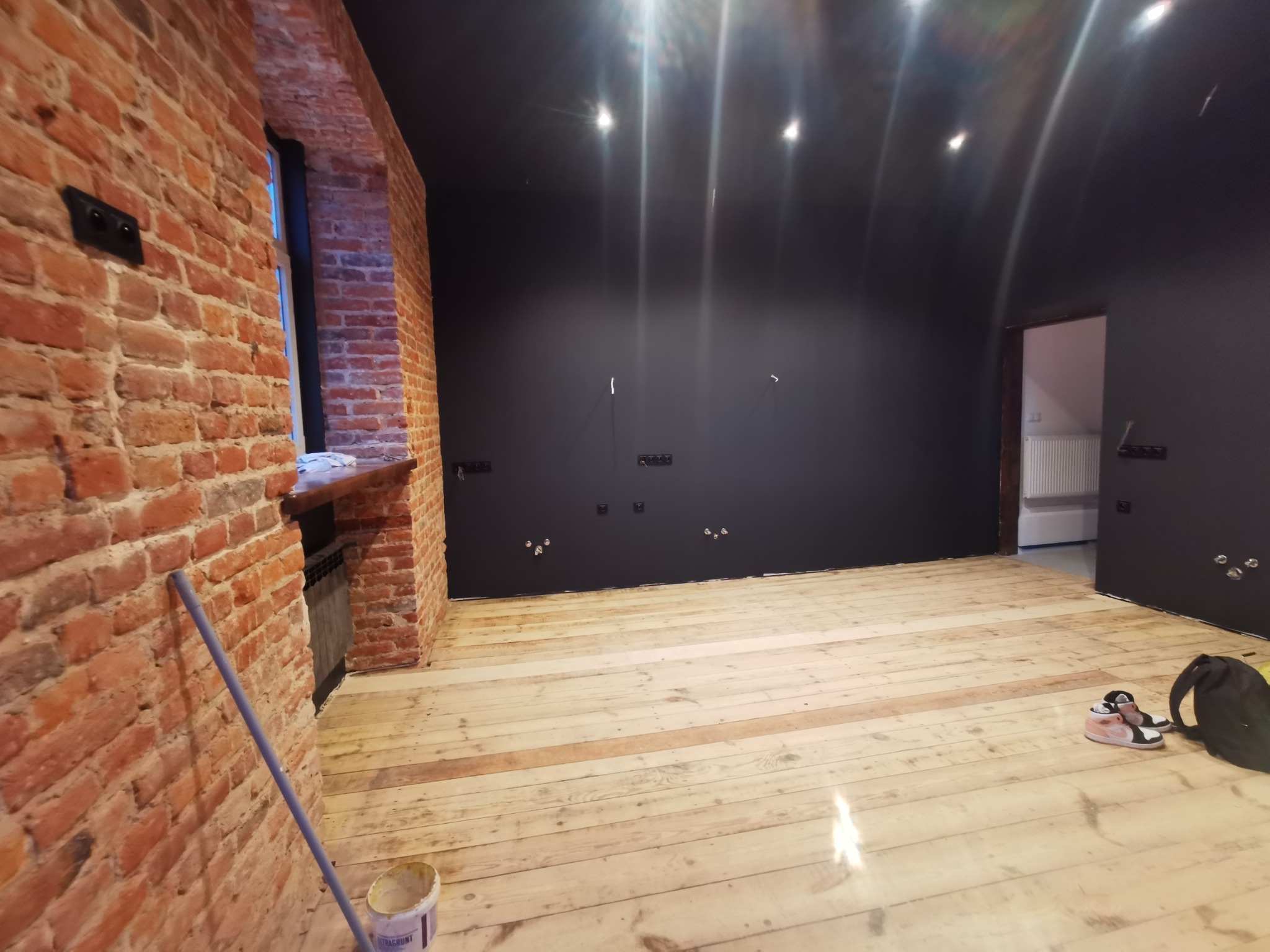 Surowe drewniane deski podłogowe w pokoju z odsłoniętą cegłą, świeżo pomalowanymi czarnymi ścianami i sufitem, oraz widocznymi puszkami elektrycznymi bez osprzętu.
