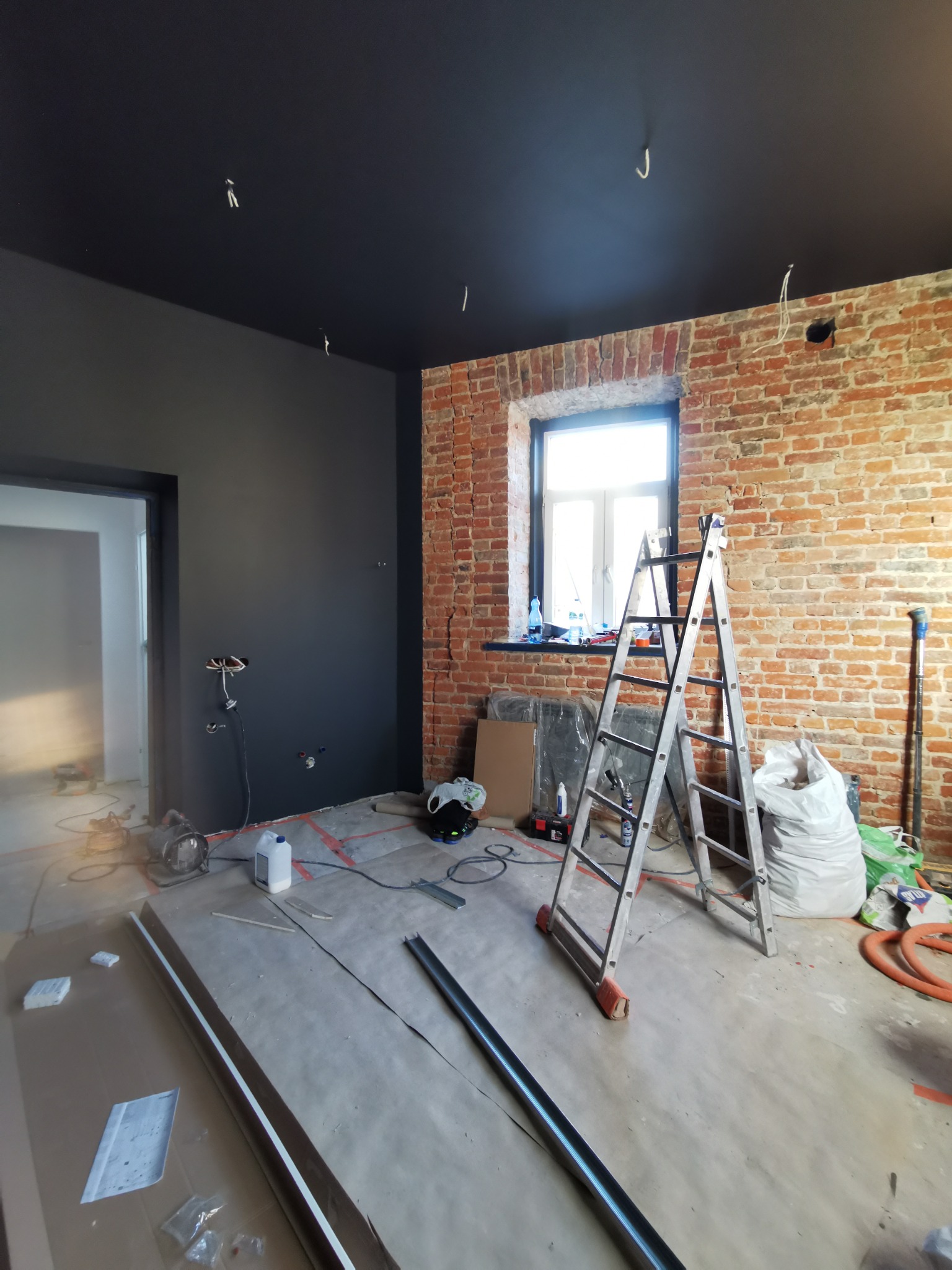 Wnętrze w trakcie remontu: pomalowany na czarno sufit, ściana z cegły, drabina aluminiowa, przygotowane materiały i narzędzia malarskie, widoczne kable elektryczne.
