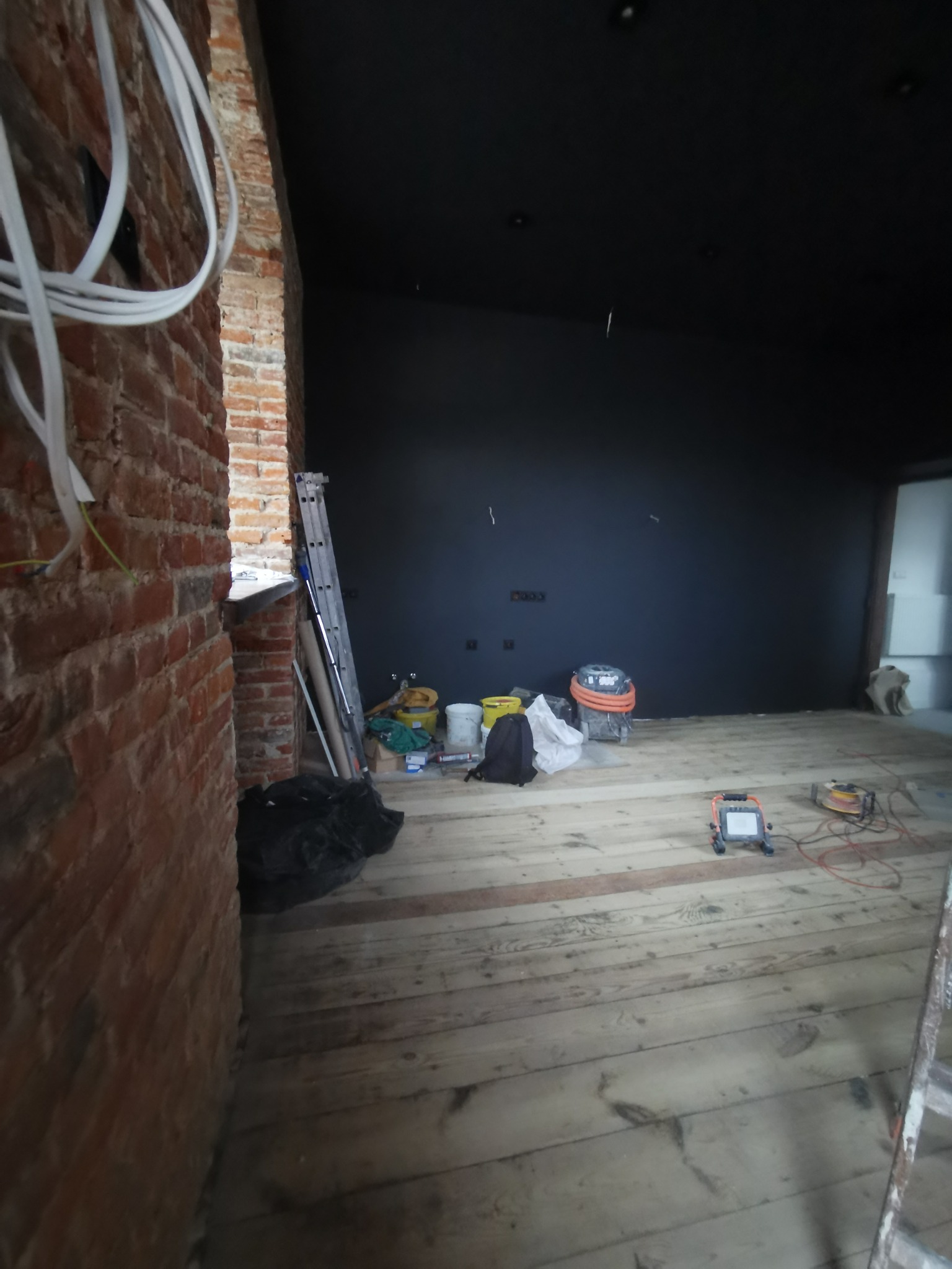 Wnętrze w trakcie remontu: surowa ceglana ściana z widocznymi przewodami, czarna ściana pomalowana na matowo, drewniana podłoga, drabina oparta o ścianę, sprzęt budowlany i wiadro z farbą w tle...