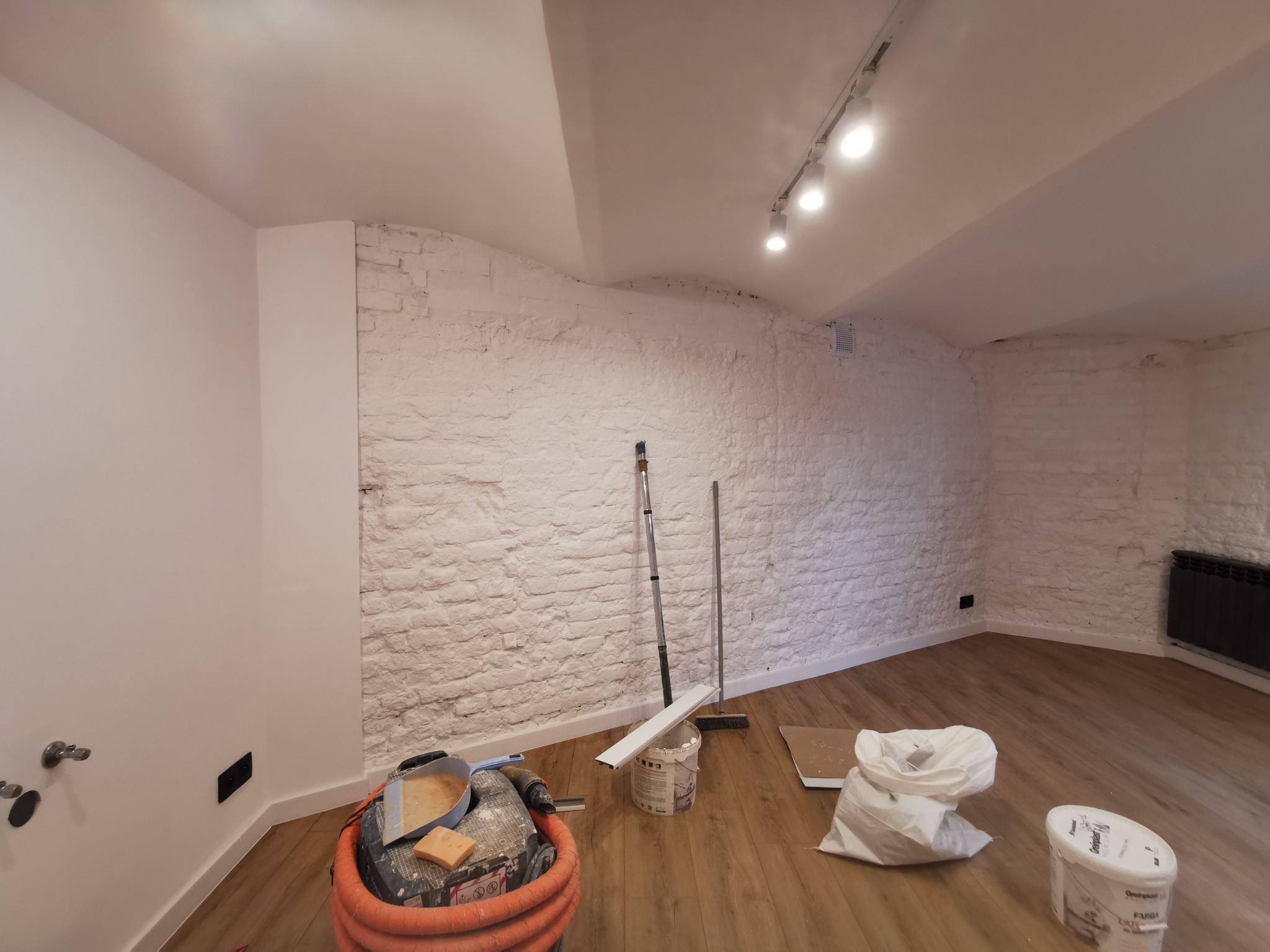 Pokój po malowaniu, z pomalowaną na biało ceglaną ścianą, widoczne narzędzia malarskie, wiadro z farbą, odkurzacz i worki z materiałami budowlanymi na podłodze.