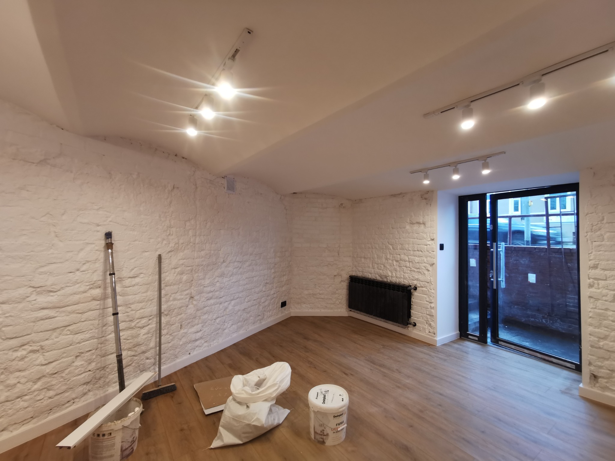 Wnętrze po odświeżeniu: biała, ceglana ściana, panele podłogowe, czarny grzejnik, przesuwne drzwi balkonowe, widoczne narzędzia malarskie i wiadro z farbą.