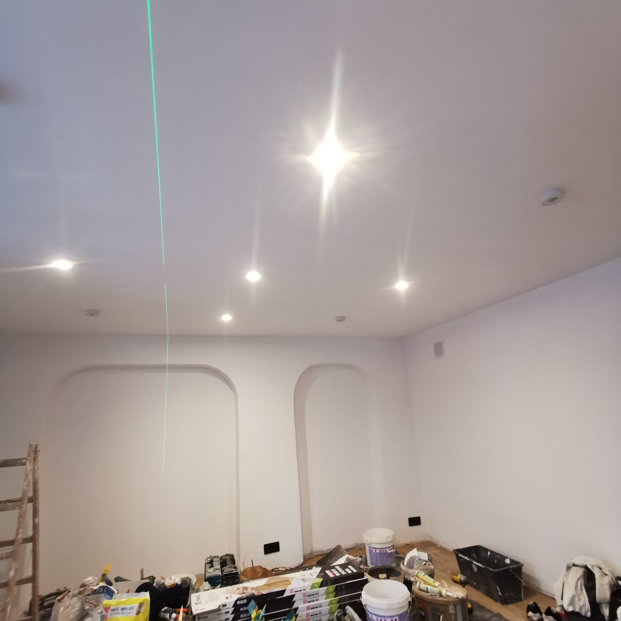 Wnętrze w trakcie prac wykończeniowych z widoczną zieloną linią lasera na ścianie, świeżo pomalowane ściany z wnękami łukowymi, widoczne narzędzia i materiały malarskie na podłodze.