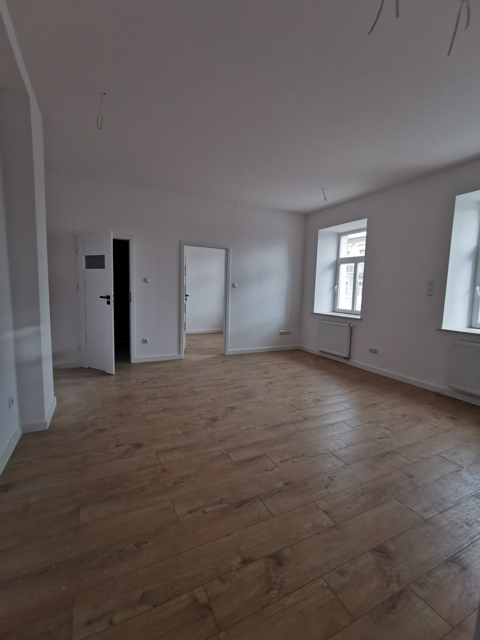 Pusty pokój po remoncie z drewnopodobną podłogą, białymi ścianami, otwartymi drzwiami i wystającymi kablami z sufitu.