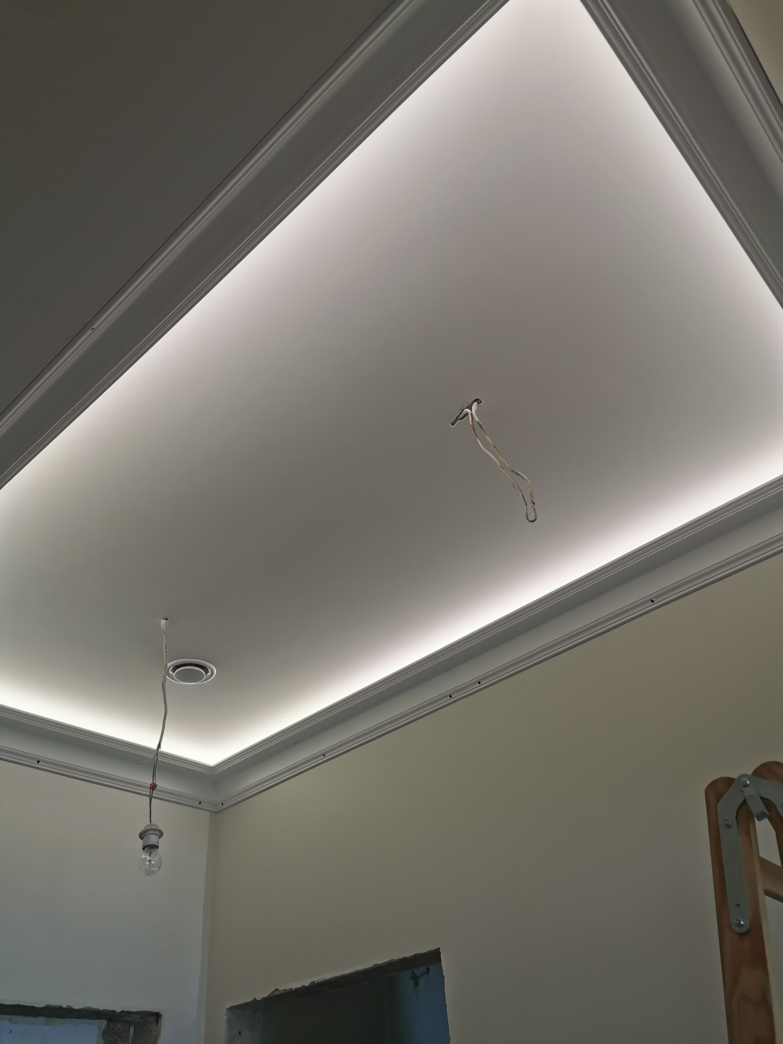 Podwieszany sufit z oświetleniem LED w trakcie montażu, widoczne wystające kable i elementy montażowe, drabina w rogu pomieszczenia.