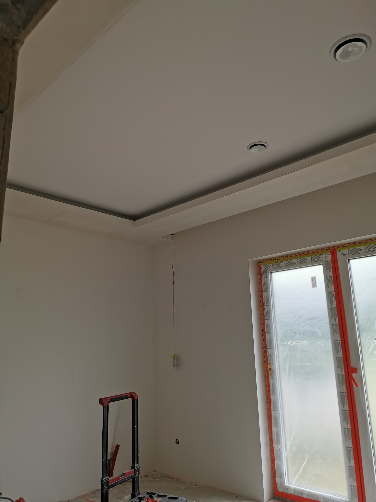 Wnętrze pokoju w trakcie wykańczania: pomalowane ściany na biało, zamontowane oświetlenie sufitowe, okno zabezpieczone folią ochronną, widoczny fragment drabiny malarskiej.