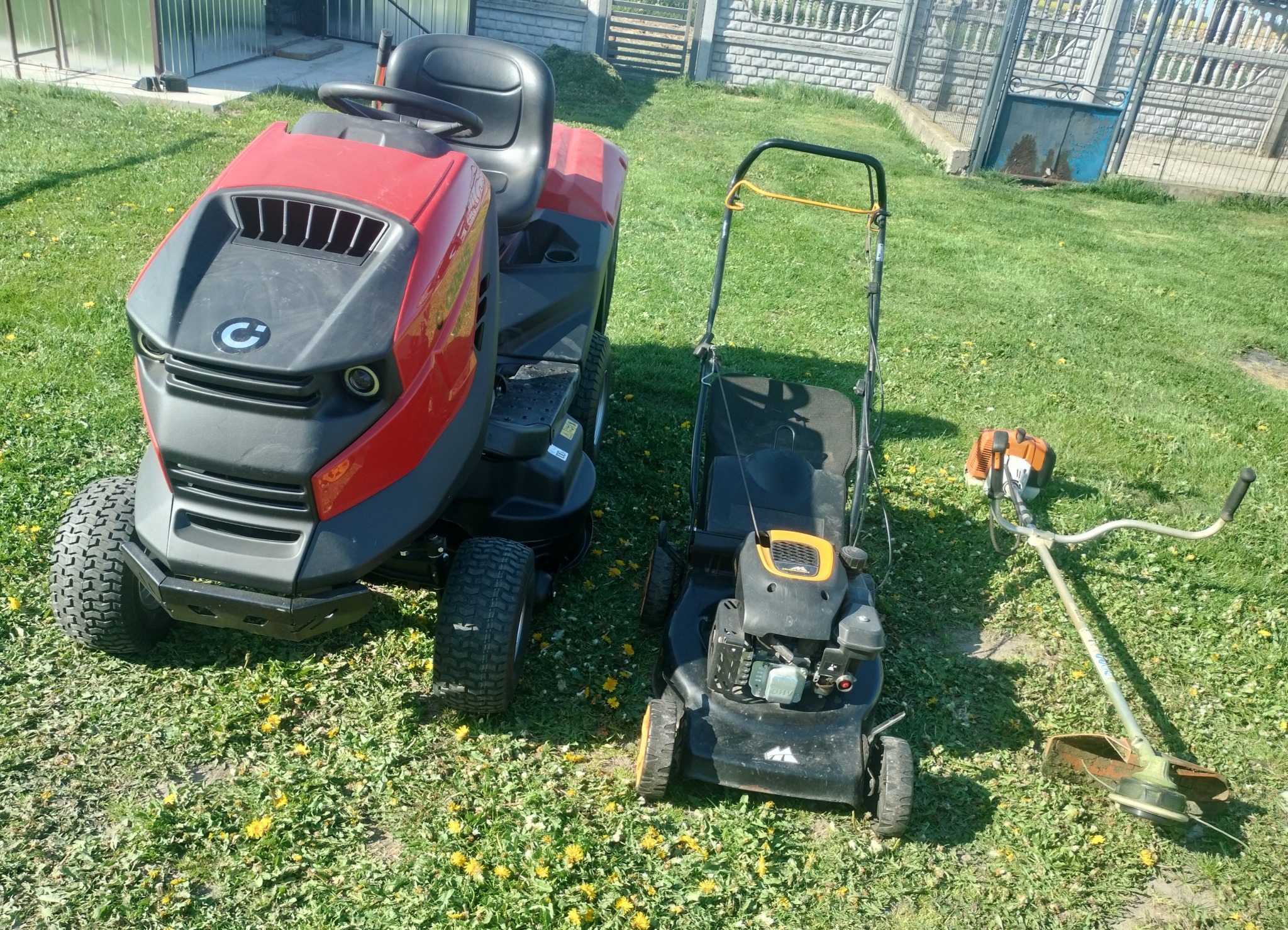 Różne typy kosiarek: traktorek, kosiarka spalinowa i podkaszarka spalinowa, stojące na trawniku z widocznymi żółtymi kwiatami, w tle ogrodzenie.