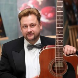 Dmytro Sokolov - Elegancki muzyk z gitarą akustyczną, ubrany w smoking i muchę, pozuje na tle obrazu z motywem owocowym. Uśmiechnięty, z gitarą w dłoniach.
