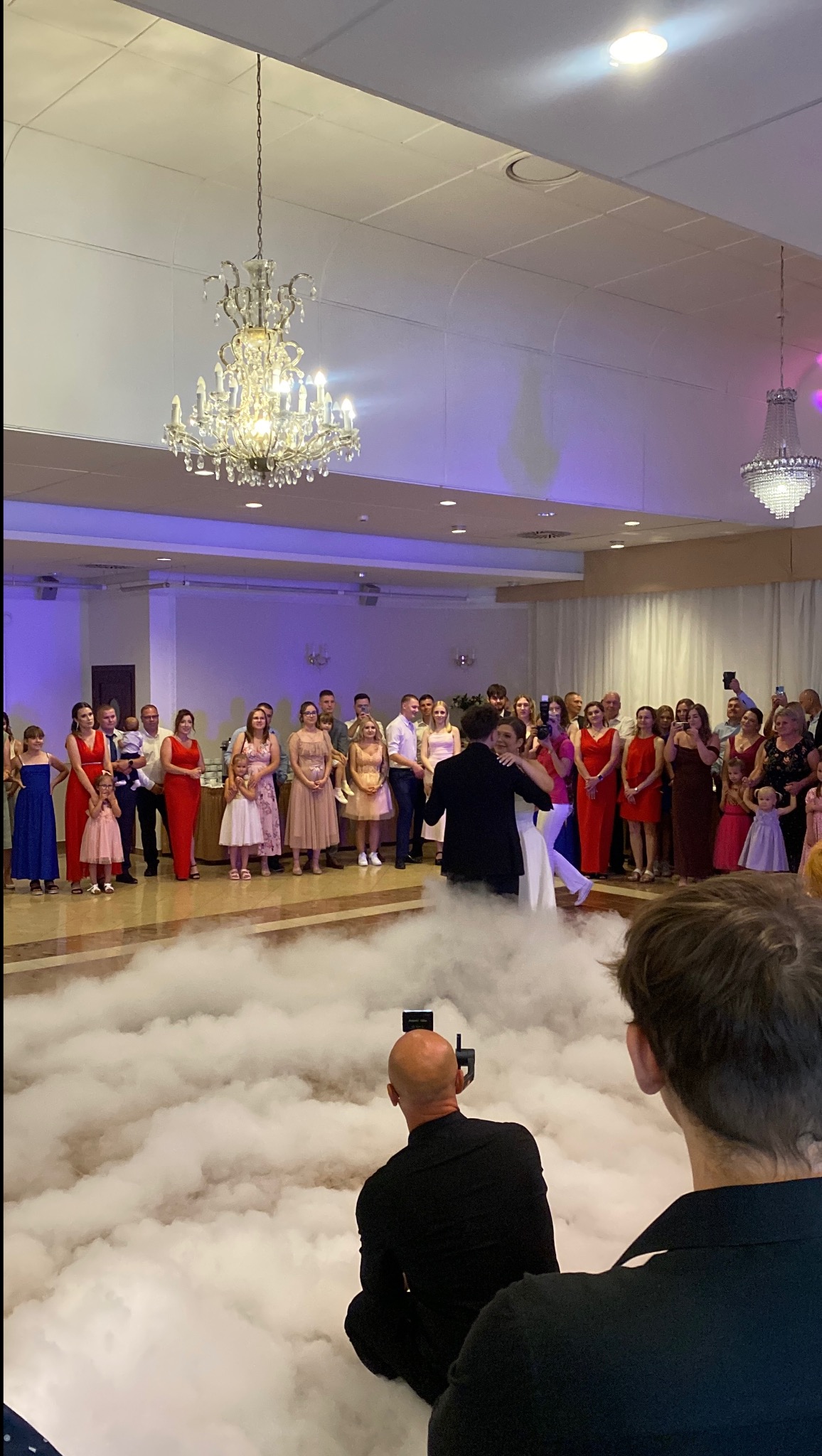 Para młoda tańczy pierwszy taniec na parkiecie wypełnionym sztuczną mgłą, otoczona gośćmi weselnymi. W tle kryształowe żyrandole i oświetlenie sceniczne.