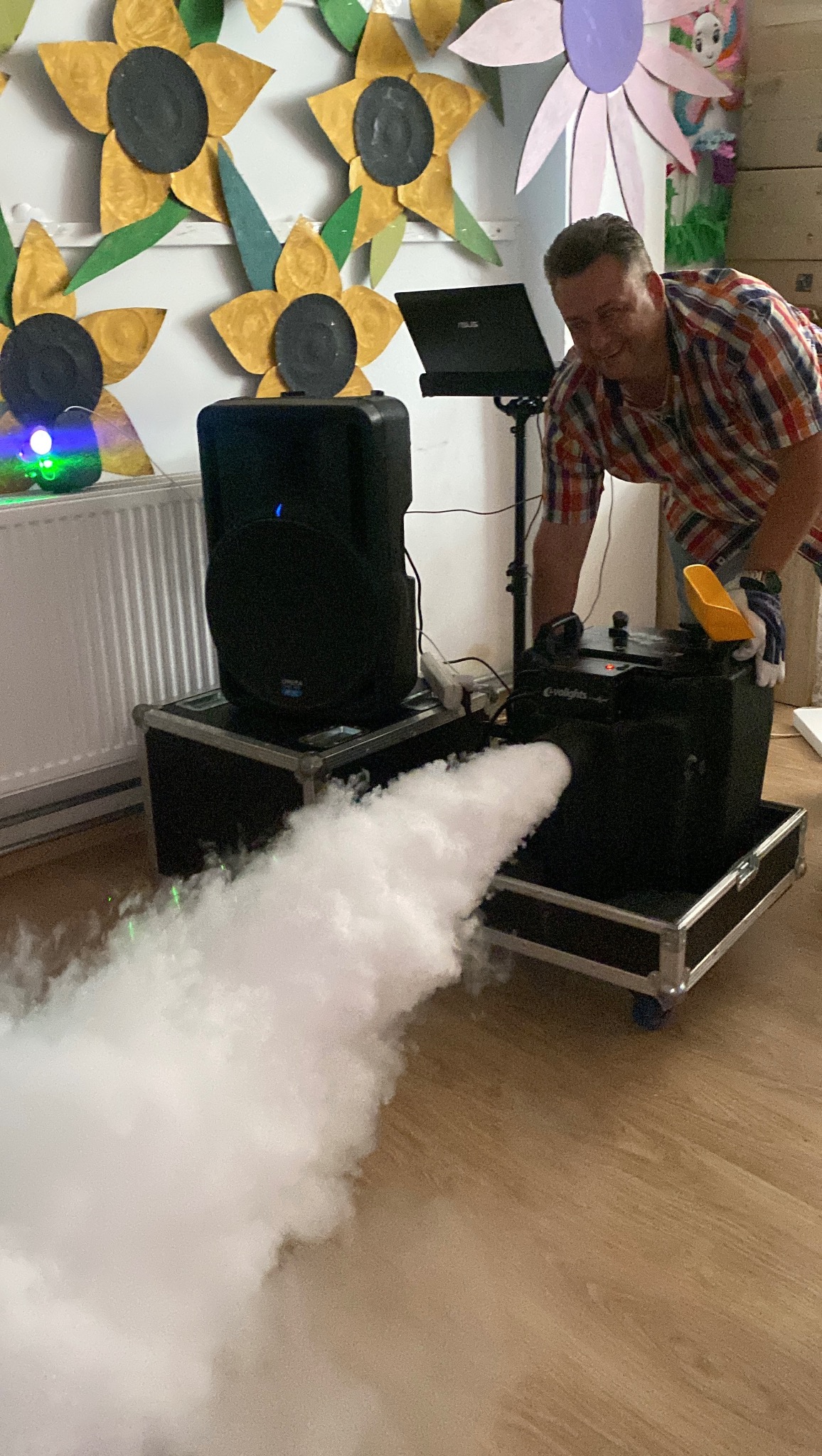 Obsługa wytwornicy dymu na imprezie: mężczyzna w koszuli w kratę kontroluje urządzenie, z którego wydobywa się gęsty dym, w tle głośniki i dekoracje.