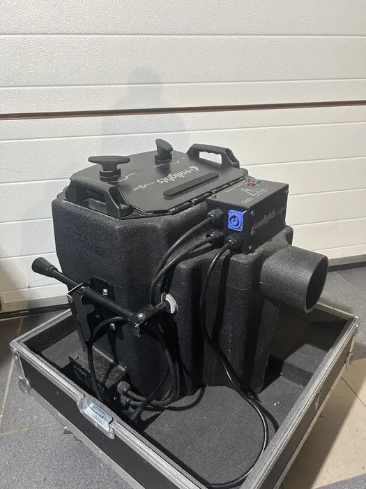 Profesjonalna wytwornica dymu Eurolite w czarnej obudowie transportowej na tle białej ściany garażowej. Widoczne przewody zasilające i elementy sterujące.