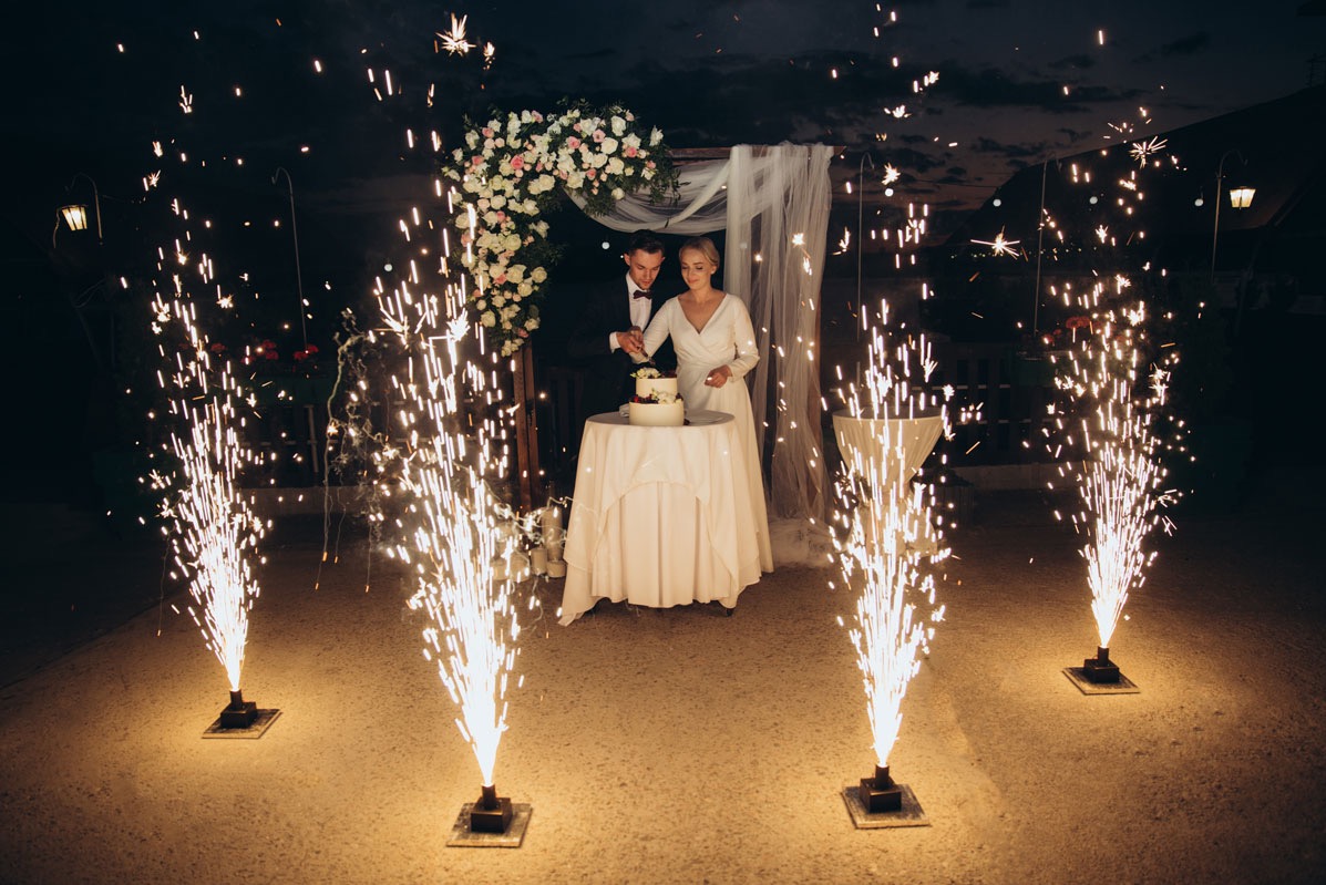 Para młoda kroi tort weselny pod dekoracyjnym łukiem z kwiatów, otoczona fontannami iskier podczas wieczornego przyjęcia.