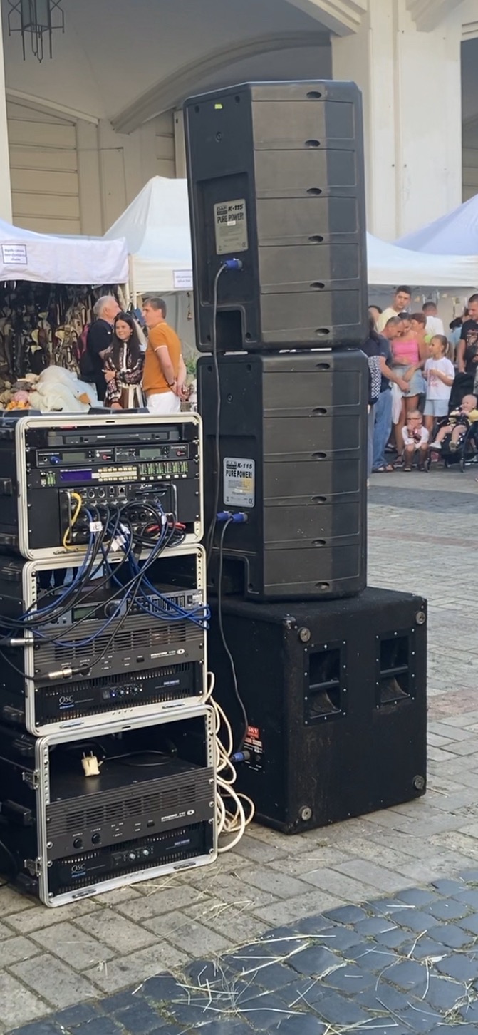 Sprzęt nagłośnieniowy: kolumny głośnikowe i rack z urządzeniami audio, z okablowaniem, na tle plenerowej sceny z publicznością i białymi namiotami.