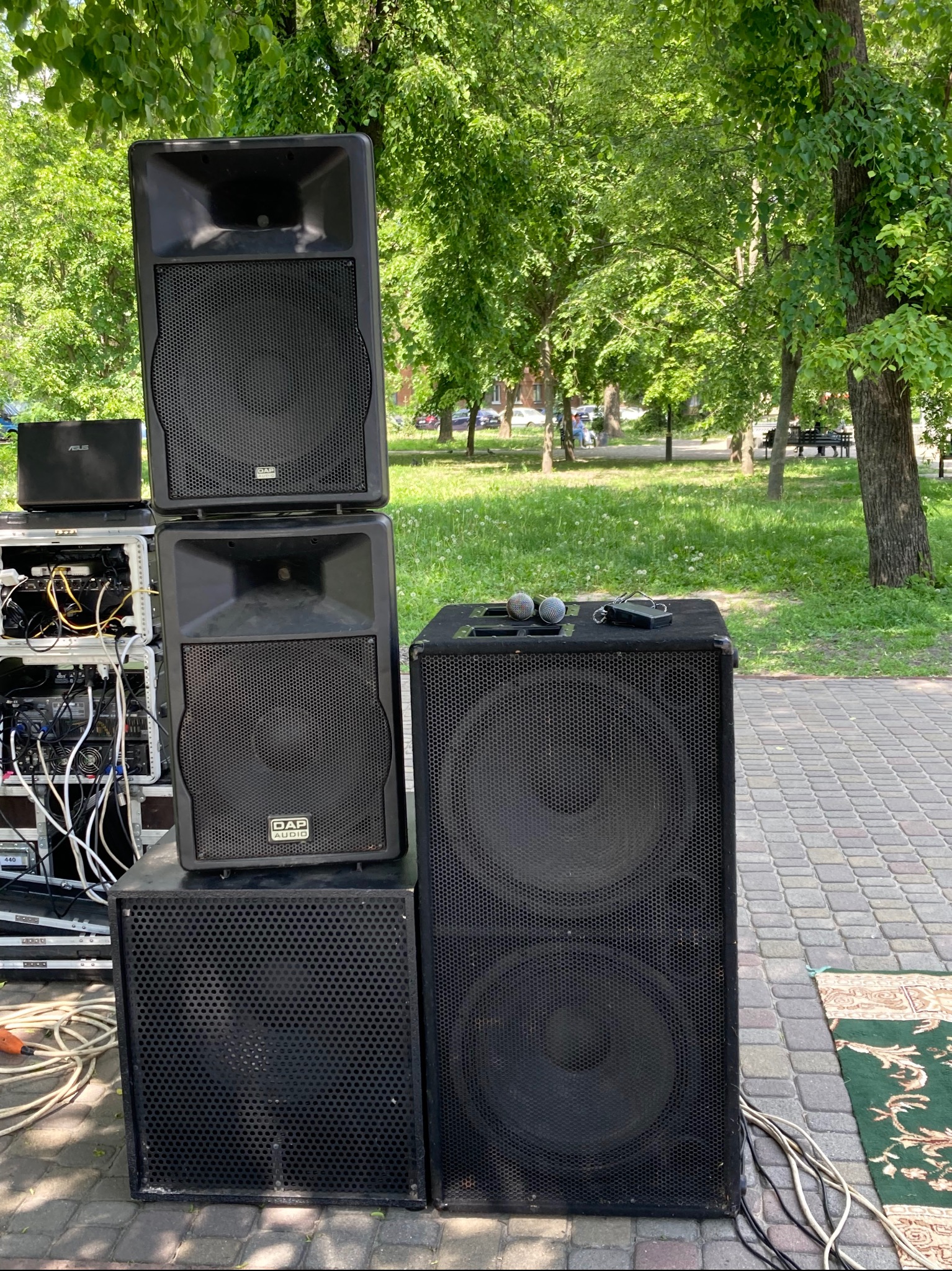 Zestaw nagłośnieniowy DAP Audio z mikrofonami na głośniku, rozstawiony na zewnątrz, prawdopodobnie przed występem zespołu muzycznego. W tle park z zielenią.