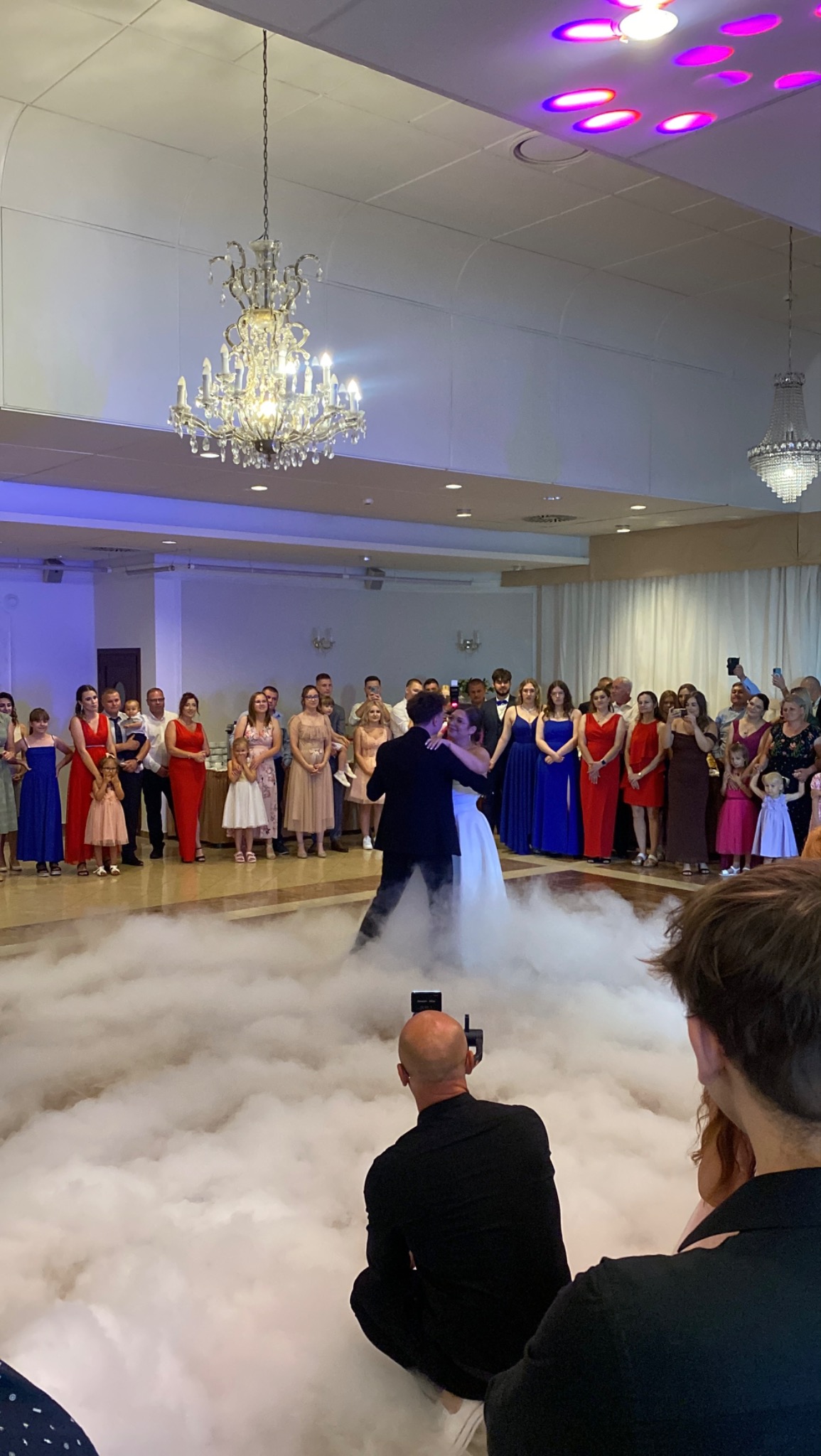 Para młoda tańczy pierwszy taniec w chmurach dymu, otoczona gośćmi weselnymi w przestronnej sali z kryształowymi żyrandolami, filmowana przez fotografa.
