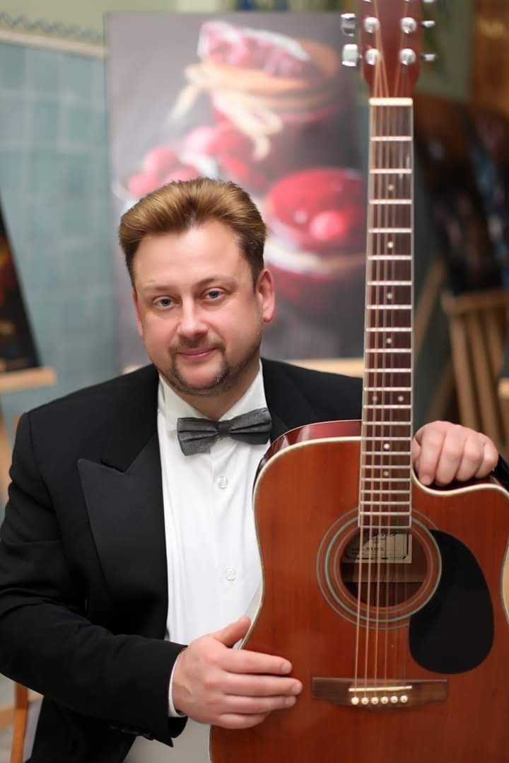 Elegancki muzyk z gitarą akustyczną, ubrany w smoking i muchę, pozuje na tle obrazu z motywem owocowym. Uśmiechnięty, z gitarą w dłoniach.