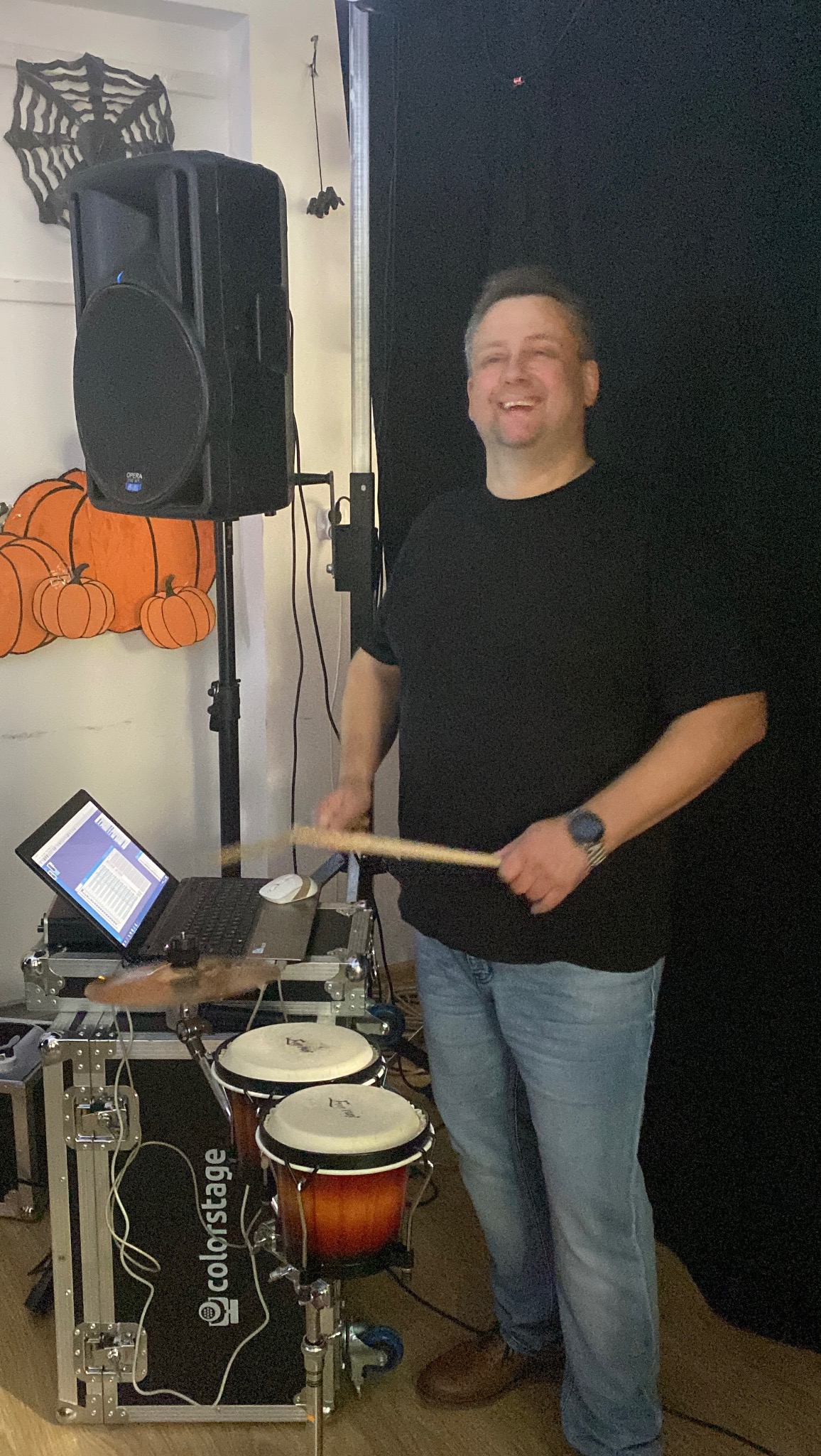 Uśmiechnięty muzyk grający na bębnach conga podczas występu, obok głośnik, laptop i dekoracje halloweenowe w tle.