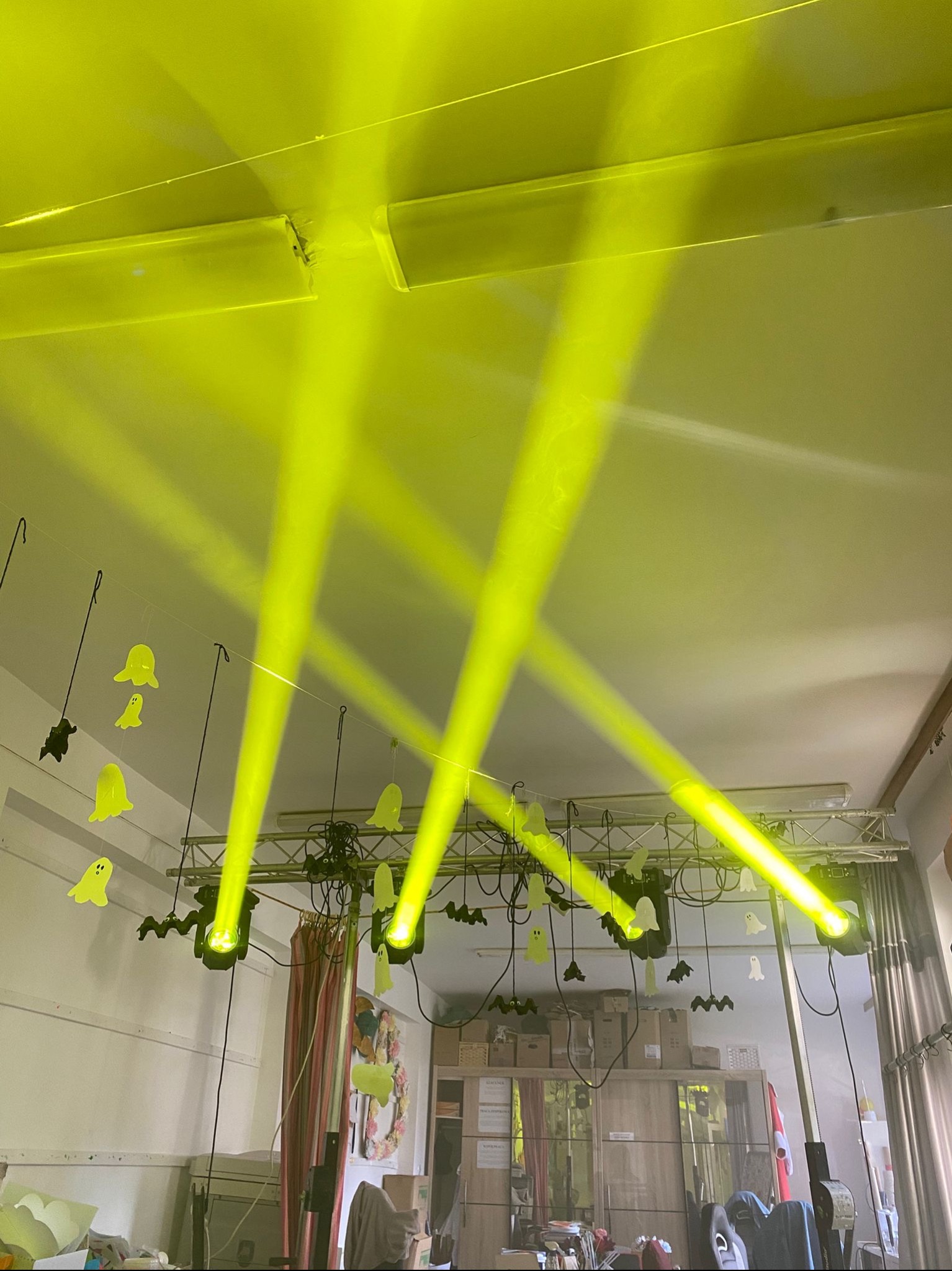 Żółte reflektory oświetlają dekoracje Halloween, w tym duchy i nietoperze, zawieszone na metalowej konstrukcji. Scena przygotowana na imprezę.