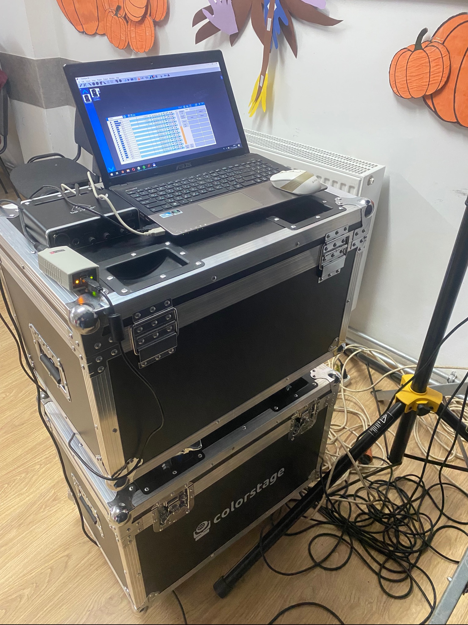 Stanowisko DJ-a z laptopem ASUS, interfejsem audio Behringer i skrzyniami transportowymi Colorstage. Na ścianie dekoracje halloweenowe.