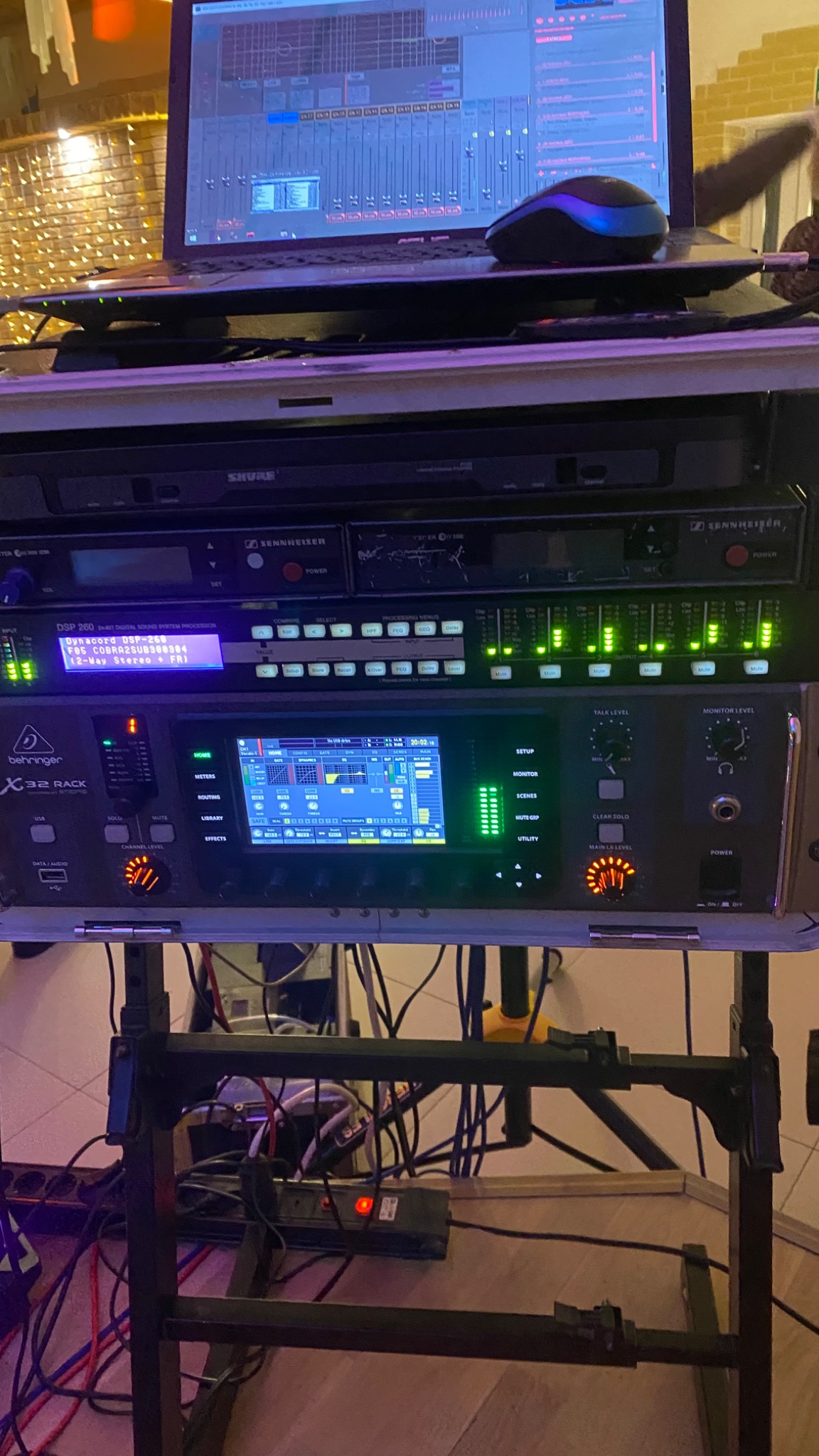 Sprzęt audio: mikser Behringer X32 Rack, procesor Dynacord DSP-268, odbiorniki Shure i Sennheiser. Laptop z oprogramowaniem audio w tle. Kable i zasilanie widoczne.