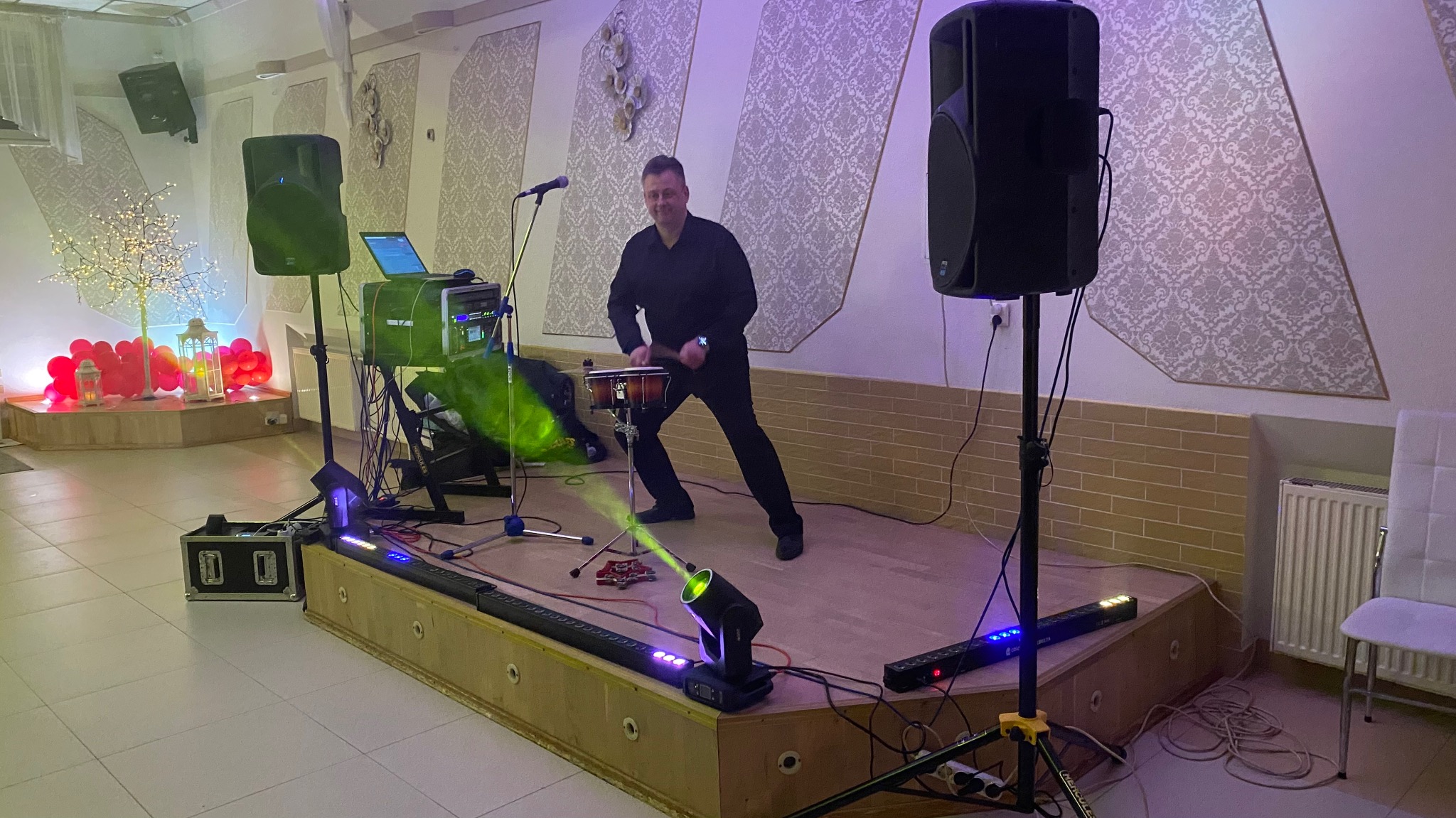 Muzyk grający na bębnach na scenie z oświetleniem i nagłośnieniem. W tle dekoracje sali.