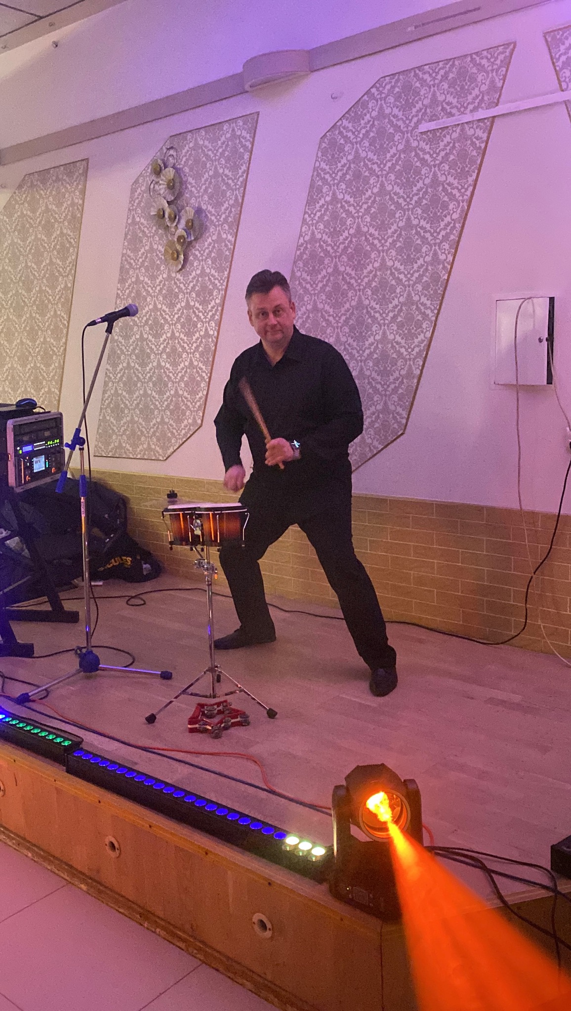 Perkusista w czarnym stroju gra na bębnie na scenie oświetlonej kolorowymi światłami. W tle mikrofon i sprzęt muzyczny.