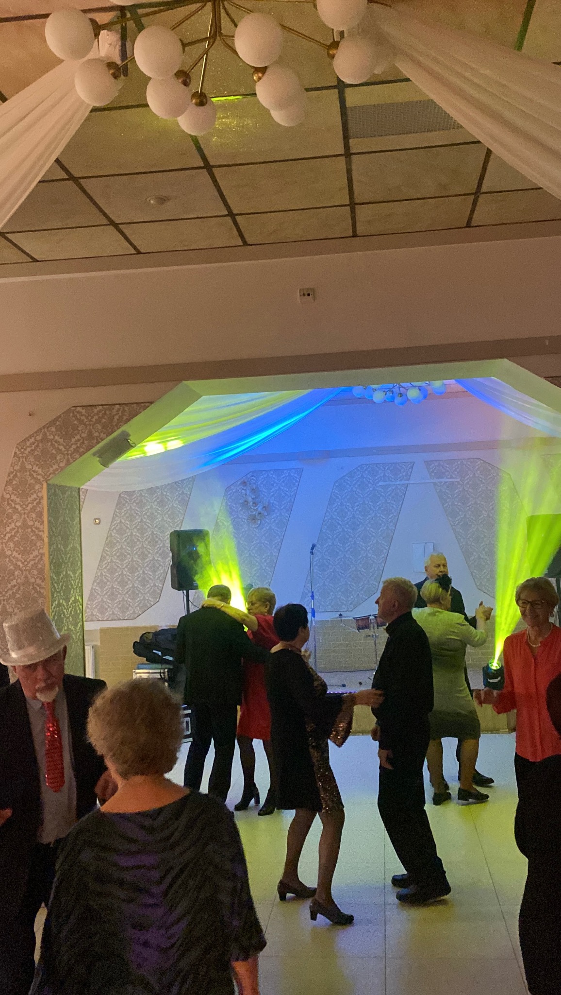 Parkiet taneczny z bawiącymi się gośćmi weselnymi, oświetlony kolorowymi reflektorami. W tle scena z instrumentami i dekoracjami. Ujęcie z perspektywy uczestnika zabawy.