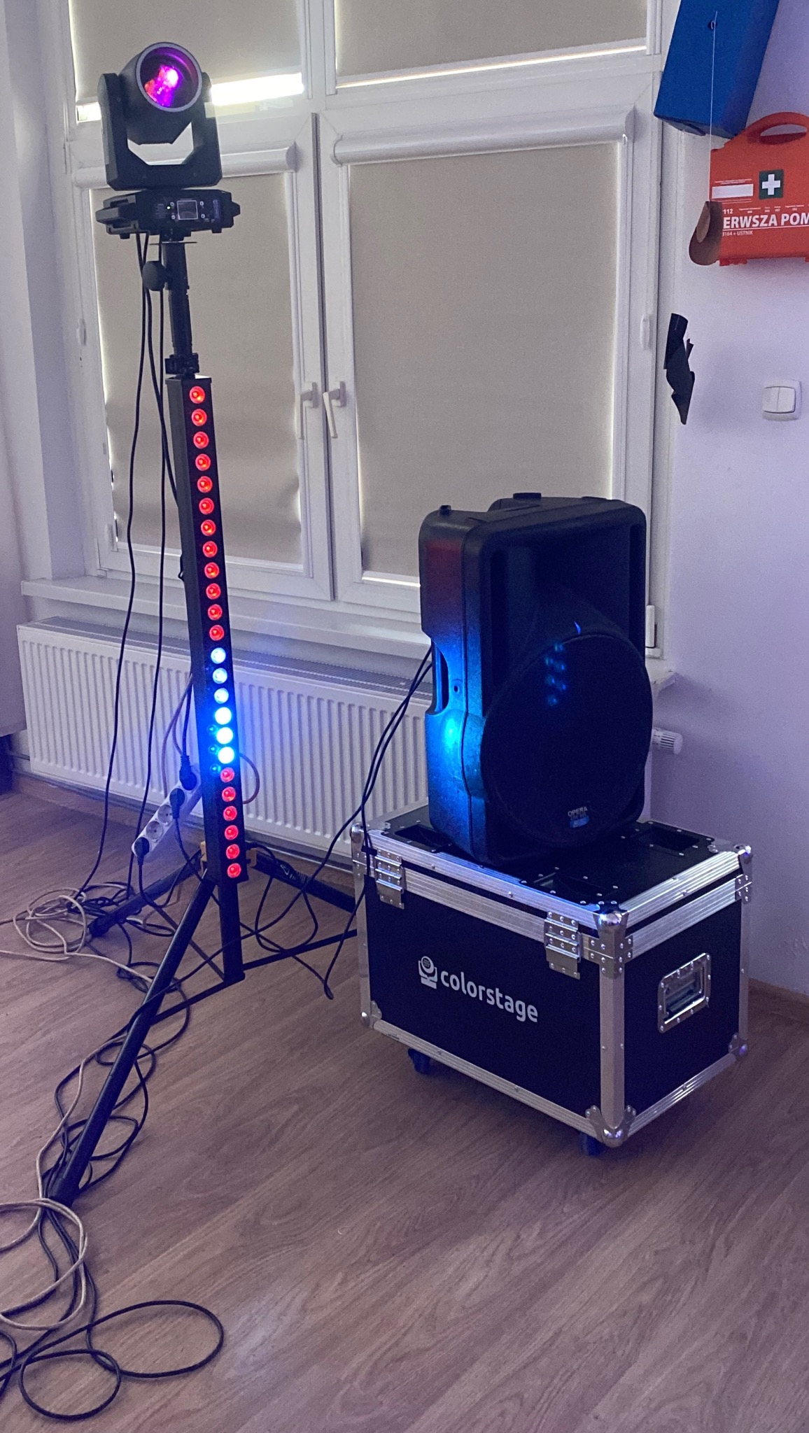Sprzęt nagłośnieniowy i oświetleniowy: kolumna głośnikowa na skrzyni Colorstage, obok listwa LED na statywie, w tle okno i apteczka pierwszej pomocy.