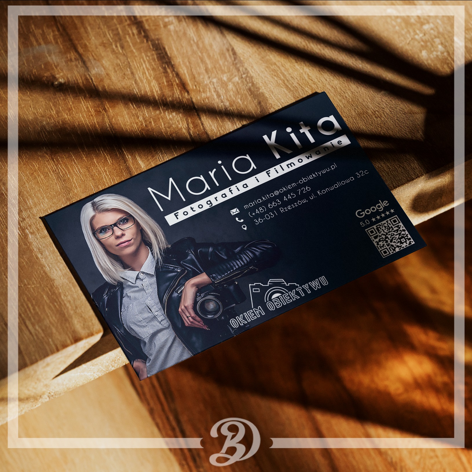 Wizytówka fotografa i filmowca Maria Kita na drewnianym tle z cieniem, prezentująca zdjęcie kobiety z aparatem, dane kontaktowe oraz logo firmy.