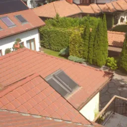 Widok z góry na dachy domów z dachówką ceramiczną, widoczne okna dachowe, panele słoneczne na jednym z dachów oraz bujna zieleń ogrodowa w tle.