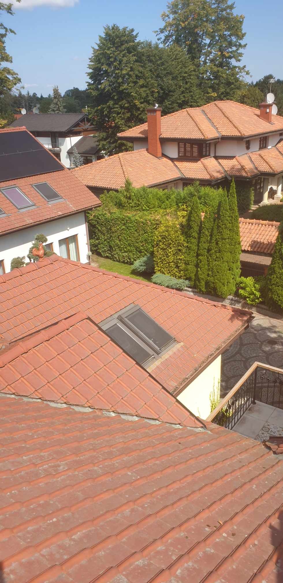 Widok z góry na dachy domów z dachówką ceramiczną, widoczne okna dachowe, panele słoneczne na jednym z dachów oraz bujna zieleń ogrodowa w tle.