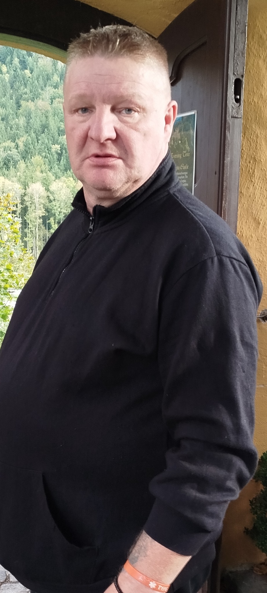 Mężczyzna w czarnej bluzie z zamkiem, stojący przy wejściu z widokiem na zalesione wzgórza w tle.