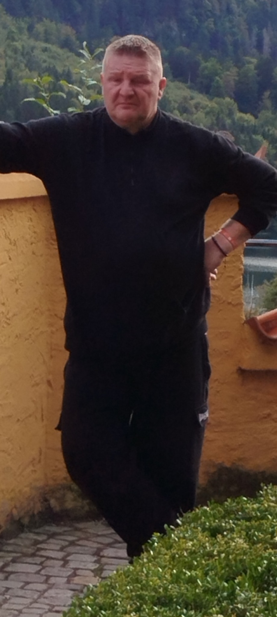 Mężczyzna w czarnym ubraniu oparty o pomarańczową ścianę z fakturą, w tle zielone wzgórza i woda.